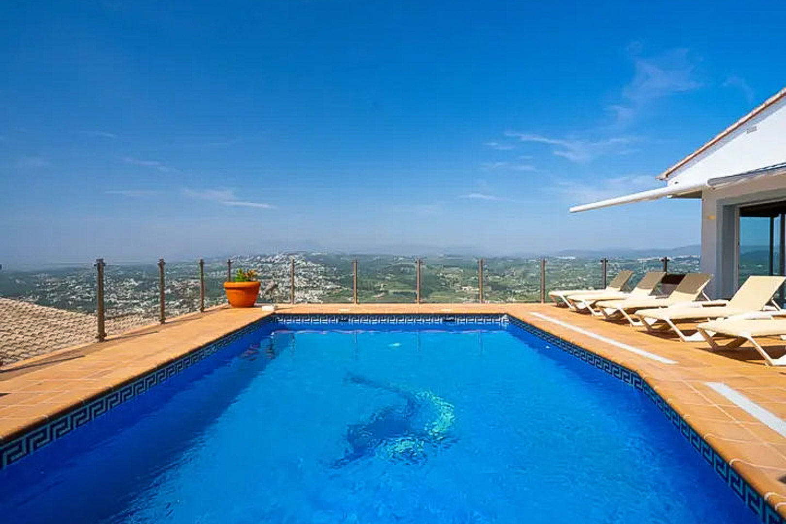 Piscine rectangulaire avec mosaïque de dauphin, terrasse ensoleillée avec transats et garde-corps en verre offrant une vue panoramique sur la vallée.