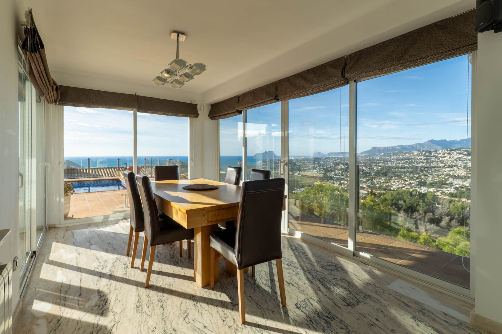 Salle à manger lumineuse avec sol en marbre et table en bois massif. De larges baies vitrées offrent une vue panoramique sur la mer Méditerranée, le Peñón de Ifach et les montagnes.
