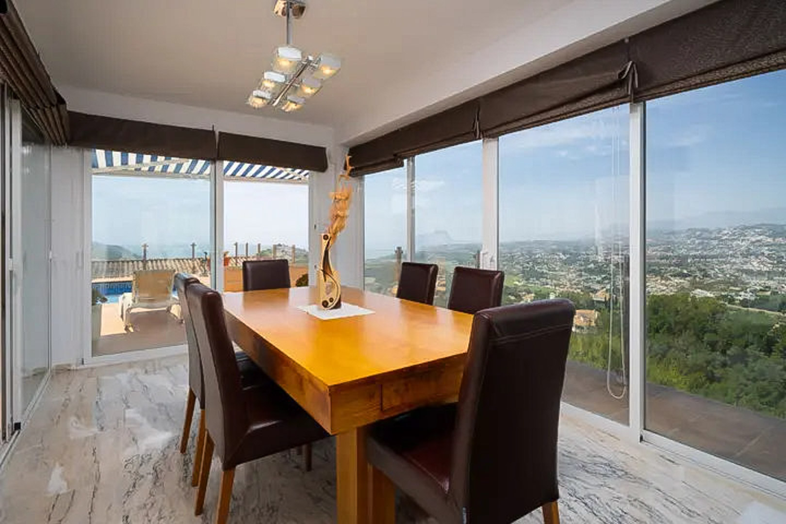 Salle à manger lumineuse avec table en bois massif et chaises en cuir, entourée de baies vitrées offrant une vue panoramique sur la mer et la côte.