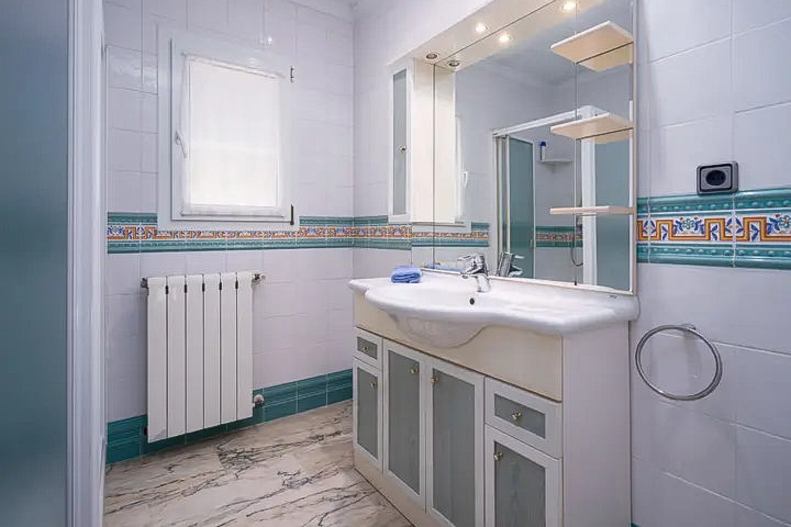 Salle de bain avec carrelage blanc et frise méditerranéenne, meuble vasque à portes en verre dépoli, grand miroir éclairé et sol en marbre veiné.