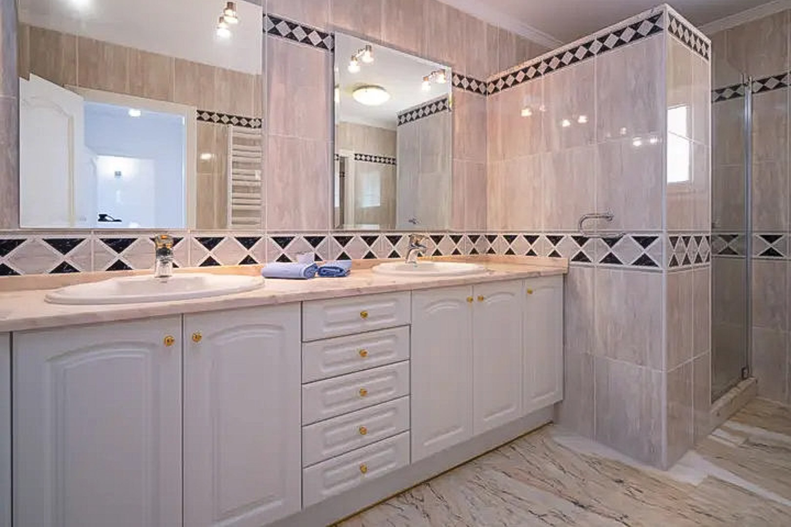 Salle de bain avec double vasque, meubles blancs classiques et plan de travail en marbre. Les murs sont ornés d'une frise en losanges noir et blanc près de la douche.
