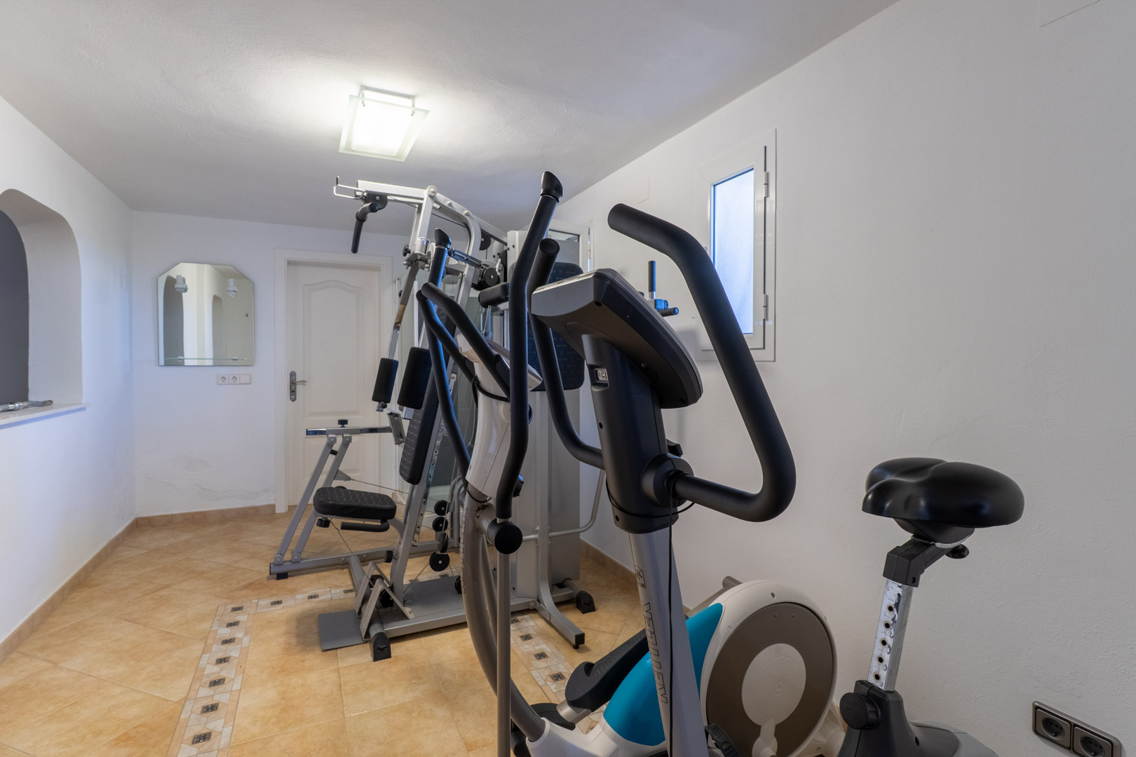 Salle de sport privée équipée d'une machine de musculation, d'un vélo elliptique et d'un vélo d'appartement sur sol carrelé.
