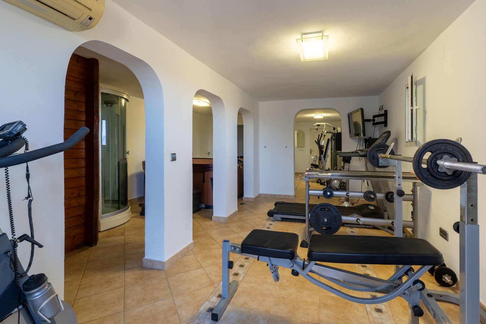 Salle de sport privée et espace bien-être avec équipements de musculation, tapis de course et accès au sauna et à la douche à vapeur par des arches blanches.