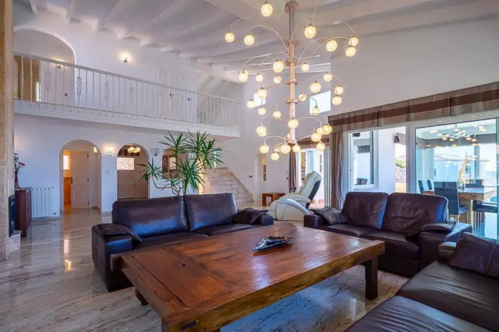 Salon à double hauteur avec canapés en cuir foncé, table basse en bois massif et lustre design. Les sols en marbre poli et la mezzanine blanche soulignent l'élégance de la pièce.