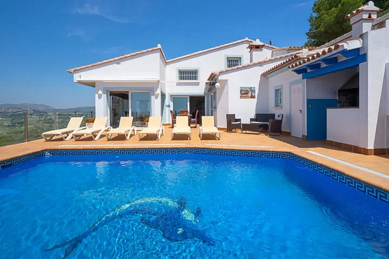Terrasse avec piscine ornée d'une mosaïque de dauphin, solarium avec transats et espace barbecue sous poutres bleues, offrant une vue dégagée.