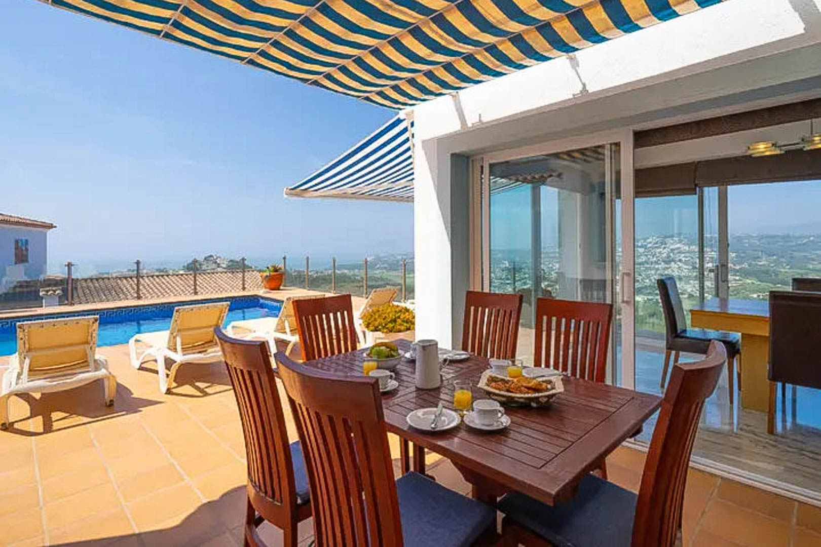Terrasse extérieure avec coin repas en bois sous un store rayé, offrant une vue sur la piscine privée et le littoral de Moraira.