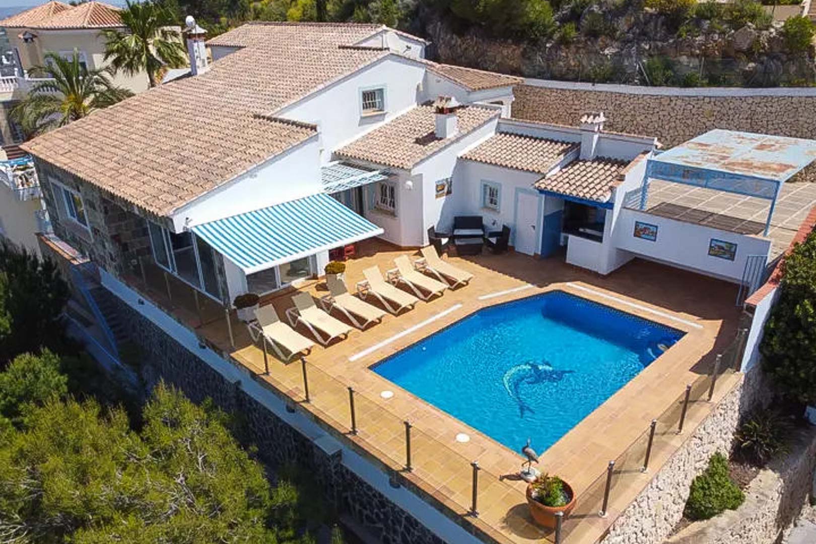 Vue aérienne de la villa présentant la piscine privée, une terrasse avec transats, un store rayé et un bar extérieur bordé de garde-corps en verre.