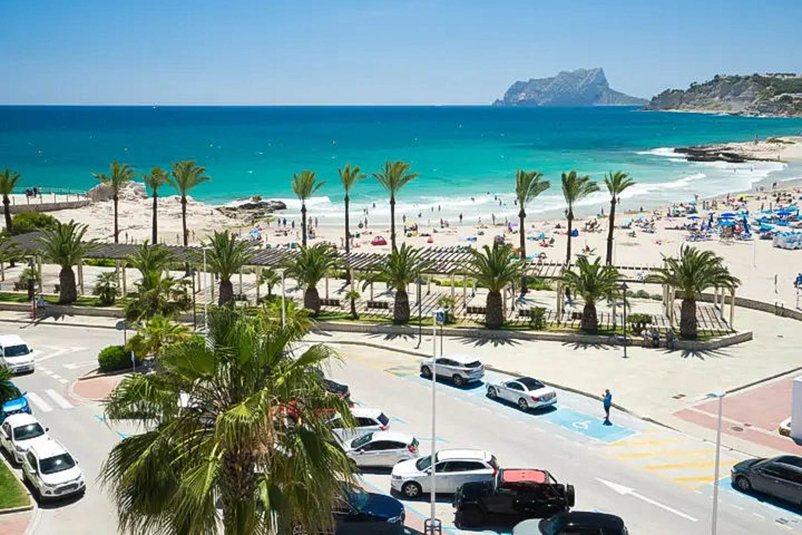 Vue panoramique de la plage de Moraira aux eaux turquoise, avec le Peñón de Ifach en arrière-plan et une promenade bordée de palmiers en premier plan.
