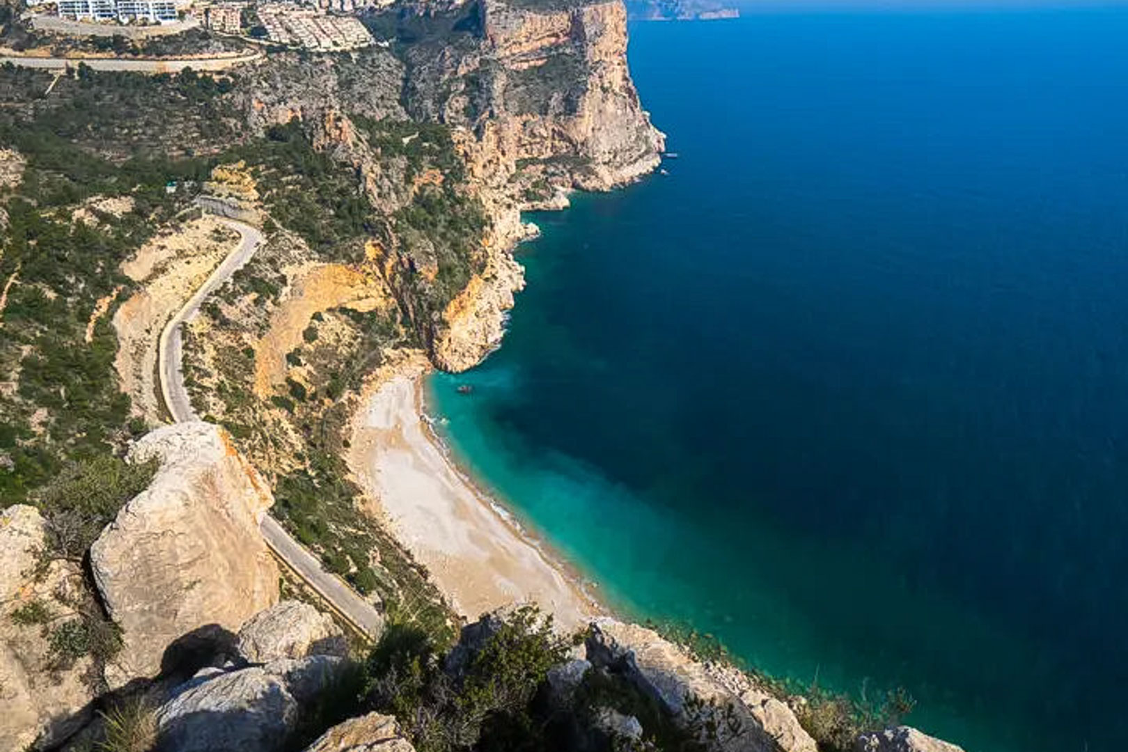 Vue panoramique sur les falaises de la Costa Blanca, avec une crique aux eaux turquoise et une route côtière sinueuse bordant la Méditerranée.