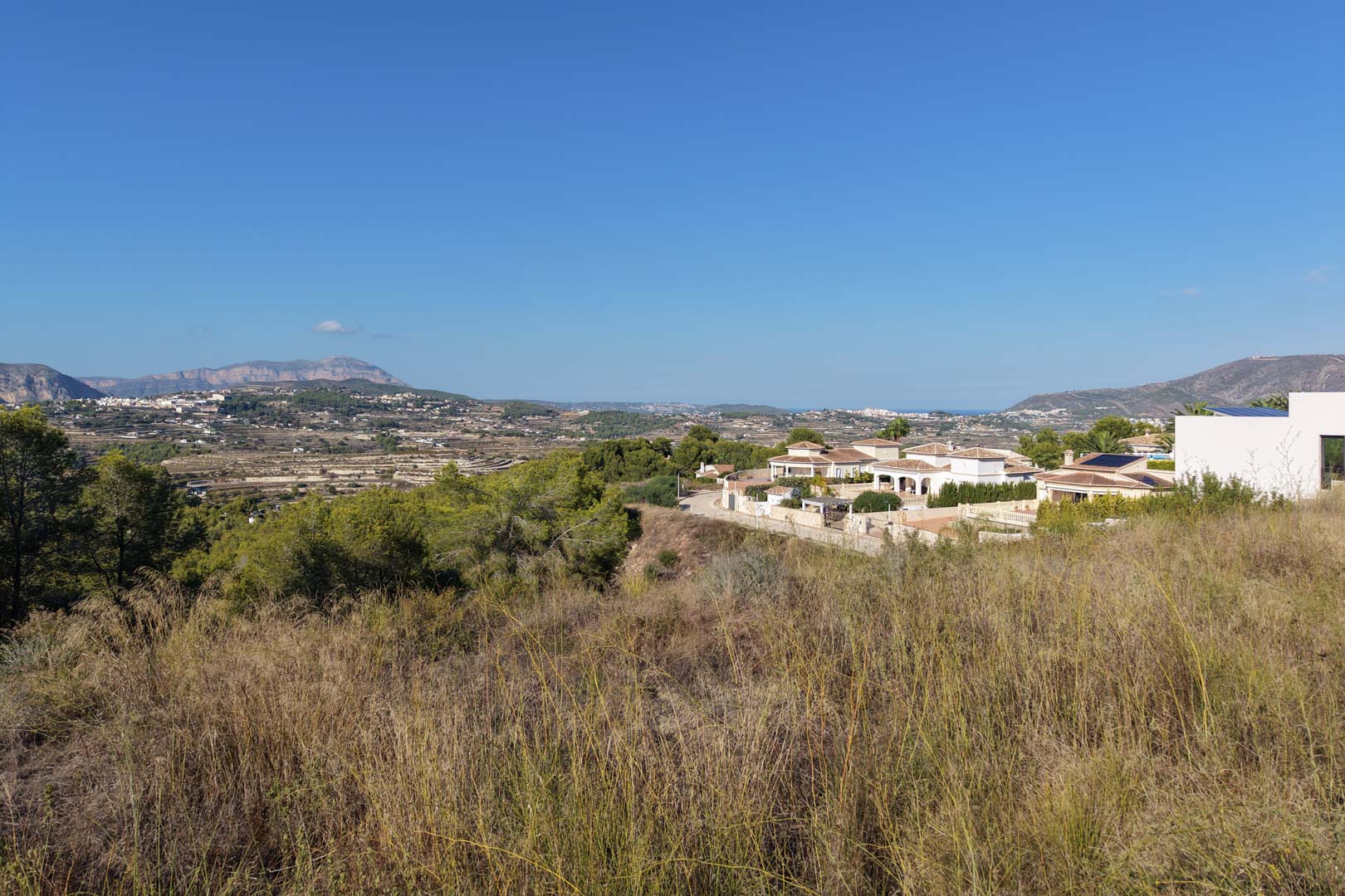 Vue surélevée du terrain urbain avec végétation sauvage, offrant des vues dégagées sur la vallée, les montagnes et les villas méditerranéennes.