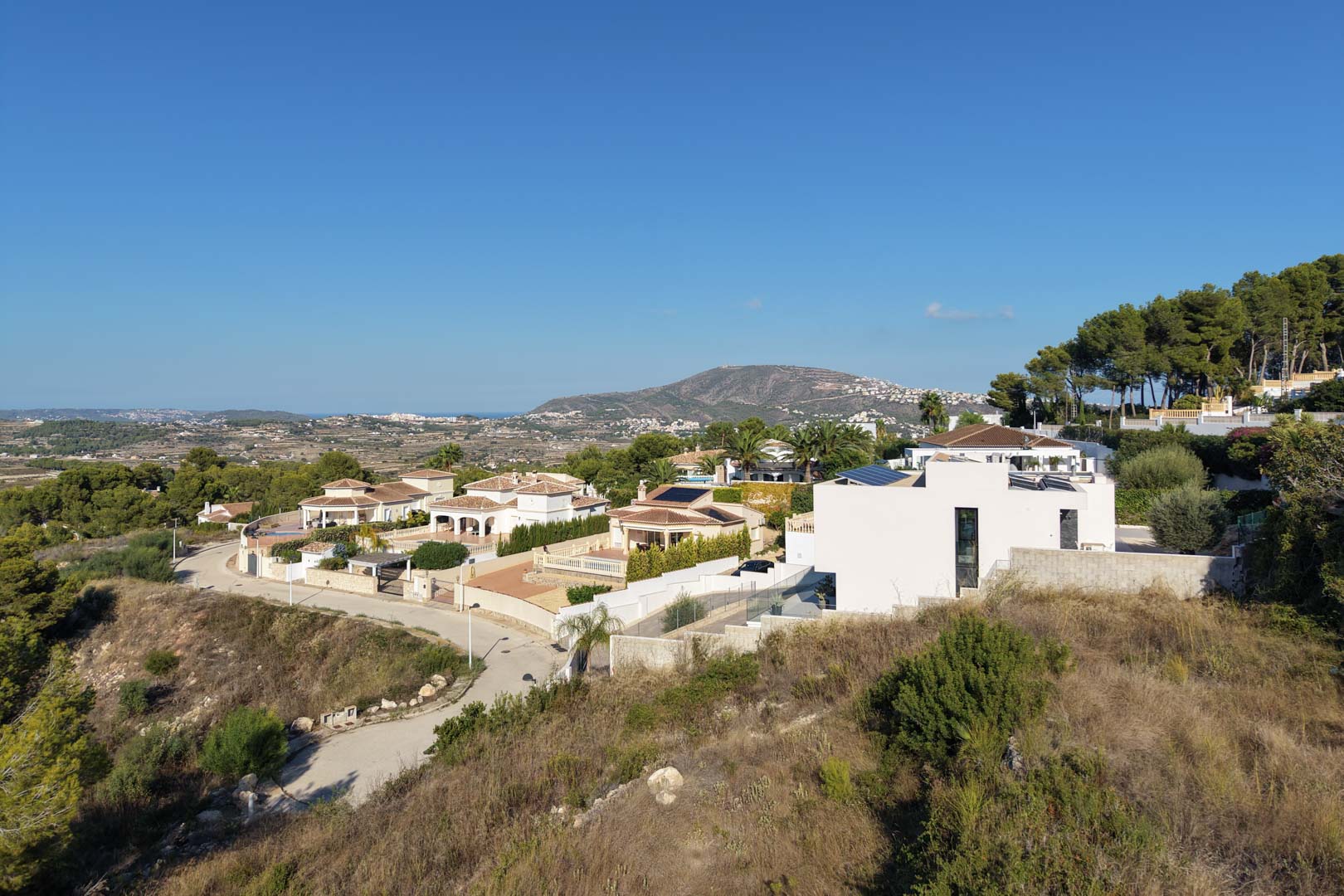 Vue panoramique surélevée du quartier résidentiel, mettant en valeur le terrain en pente et les vues dégagées vers les montagnes et la mer au loin.
