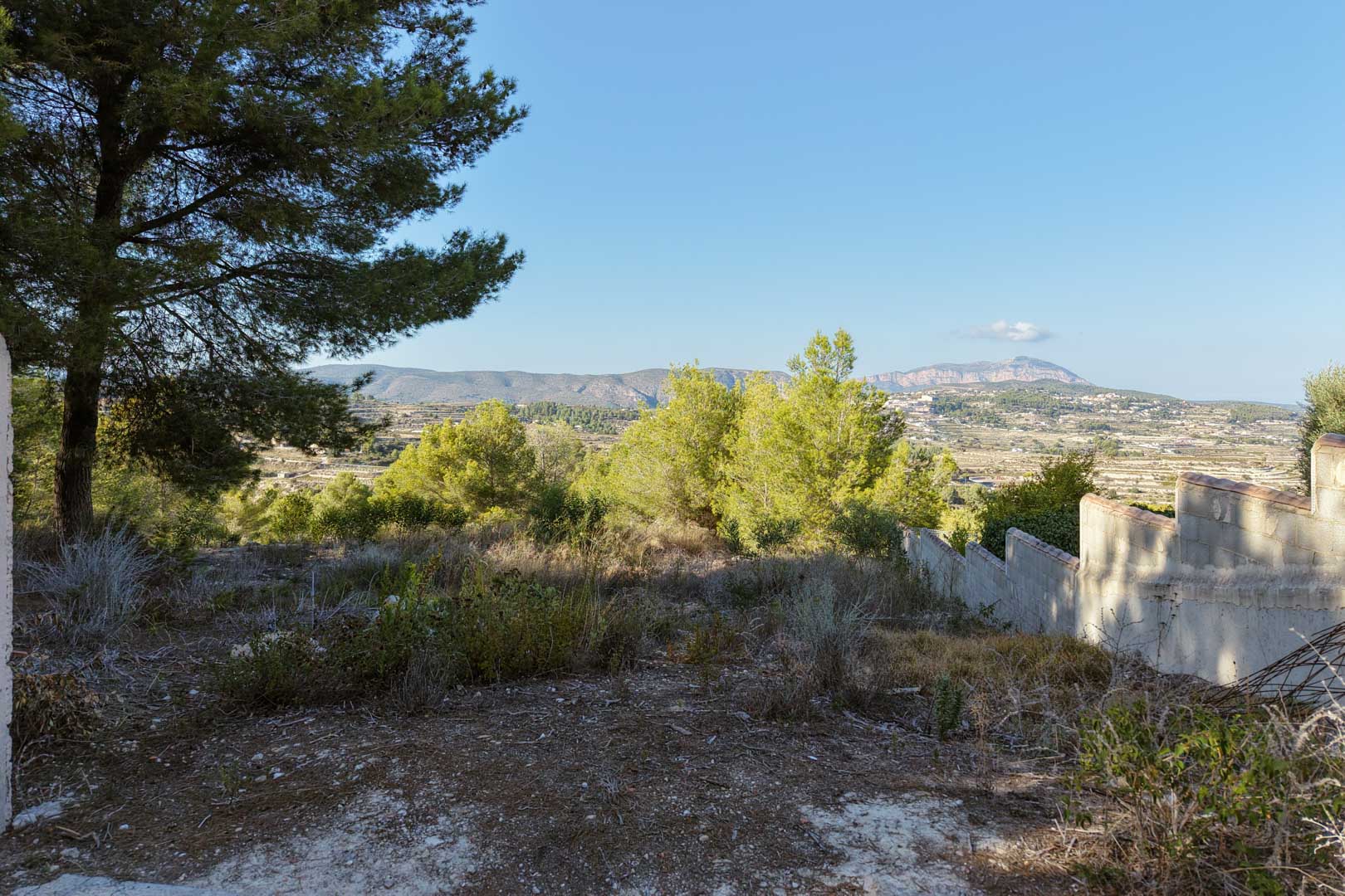 Terrain en hauteur offrant une vue panoramique sur la vallée et les montagnes. L'espace comprend une végétation naturelle, des pins et un mur de clôture en gradins.