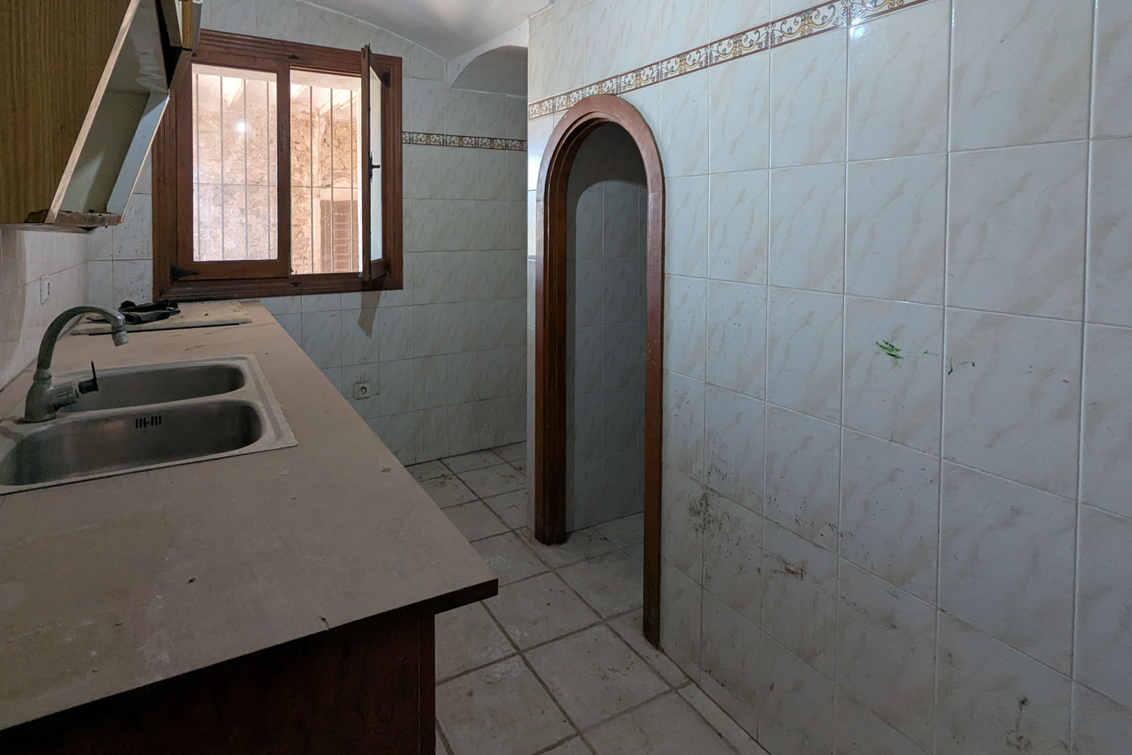 Cuisine rustique d'origine avec double évier en inox, murs carrelés avec frise décorative et une arche en bois menant aux autres pièces.
