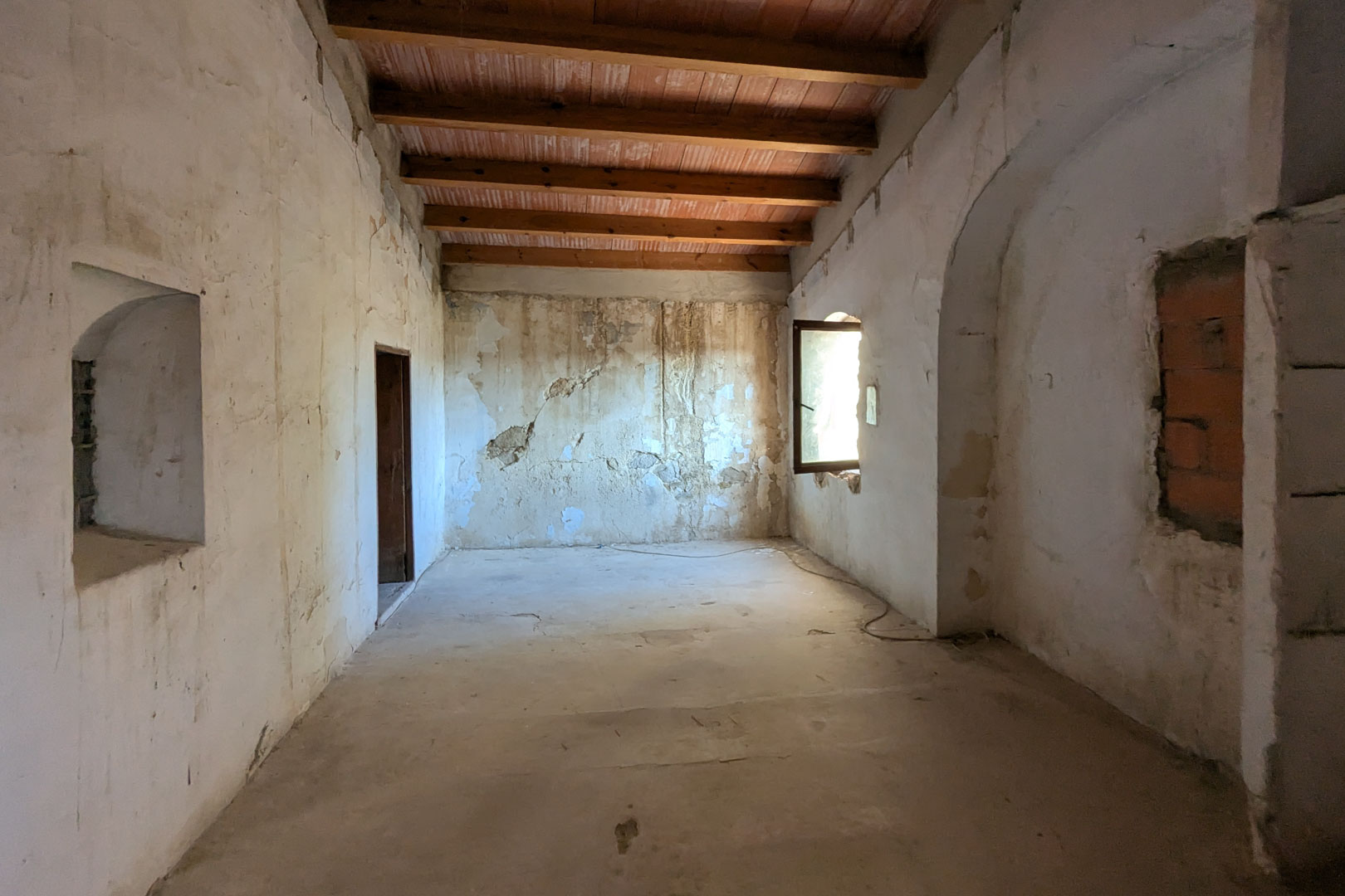 Grande pièce ouverte avec de hauts plafonds à poutres en bois et briques de terre cuite. Les murs épais présentent des niches d'origine et une fenêtre latérale lumineuse.