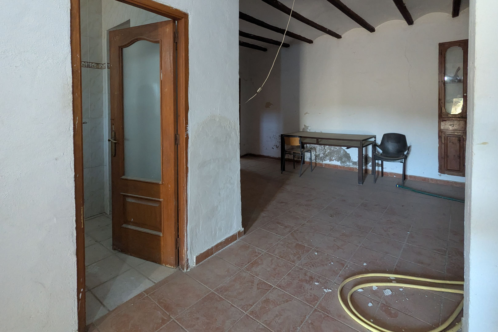 Intérieur rustique avec poutres apparentes en bois sombre, sol en terre cuite et murs à la chaux. Comprend une porte en bois vitrée et un placard mural encastré.