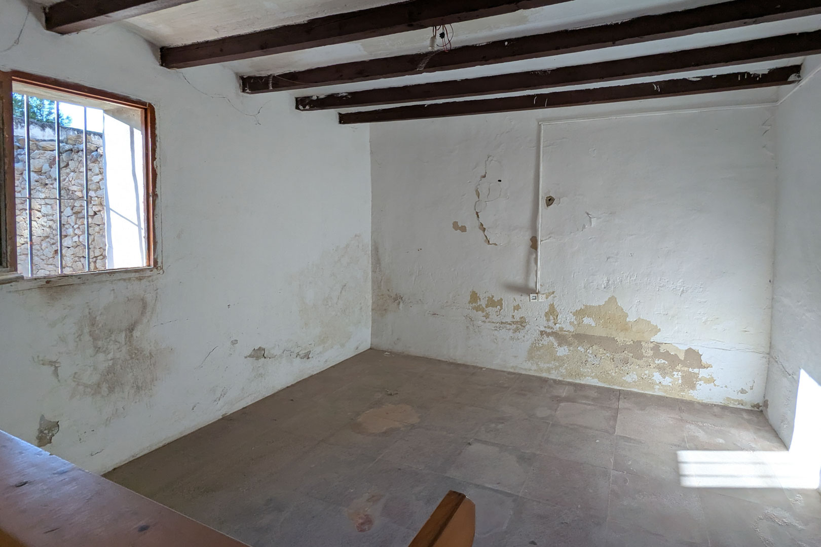Pièce rustique avec poutres apparentes en bois sombre, murs à la chaux d'origine et sol en terre cuite. Une fenêtre grillagée apporte une lumière naturelle sur les textures anciennes du bâti.
