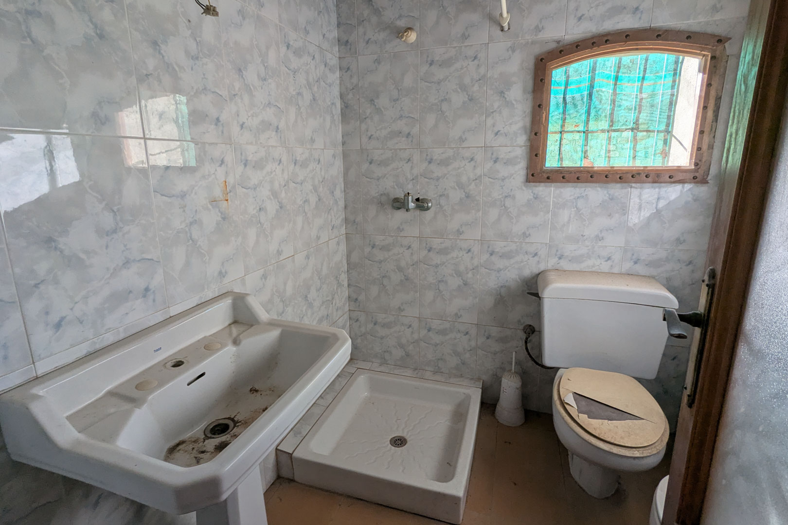 Salle de bain d'origine avec carrelage mural effet marbre bleu, lavabo sur pied, receveur de douche carré et petite fenêtre cintrée avec cadre en bois.
