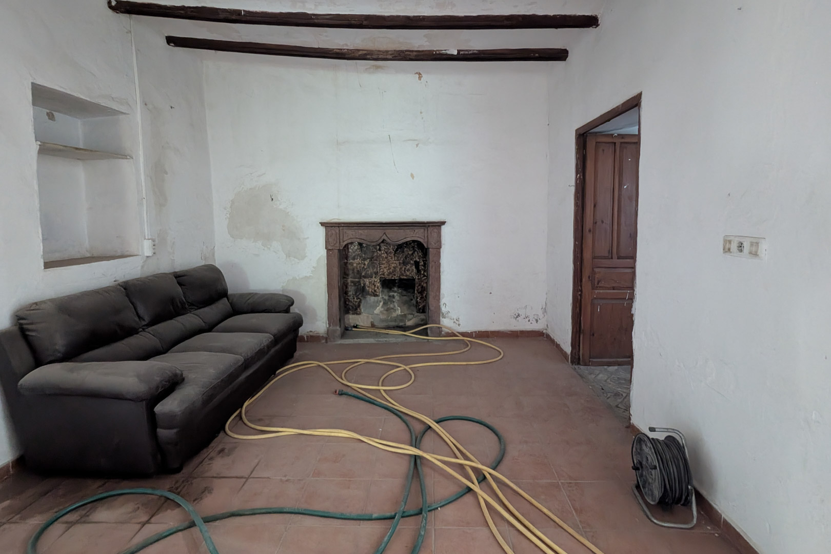 Salon traditionnel avec poutres en bois apparentes, sol en terre cuite et cheminée d'origine avec manteau en pierre sculptée. Les murs épais incluent une niche intégrée.