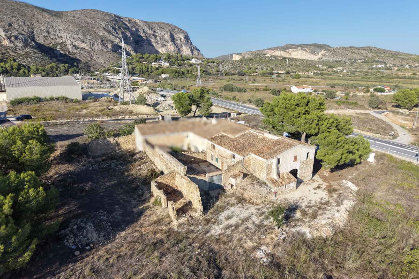 Vue aérienne de la ferme traditionnelle en pierre avec cour intérieure et annexes, située sur un vaste terrain rustique avec vue sur les montagnes de Teulada.
