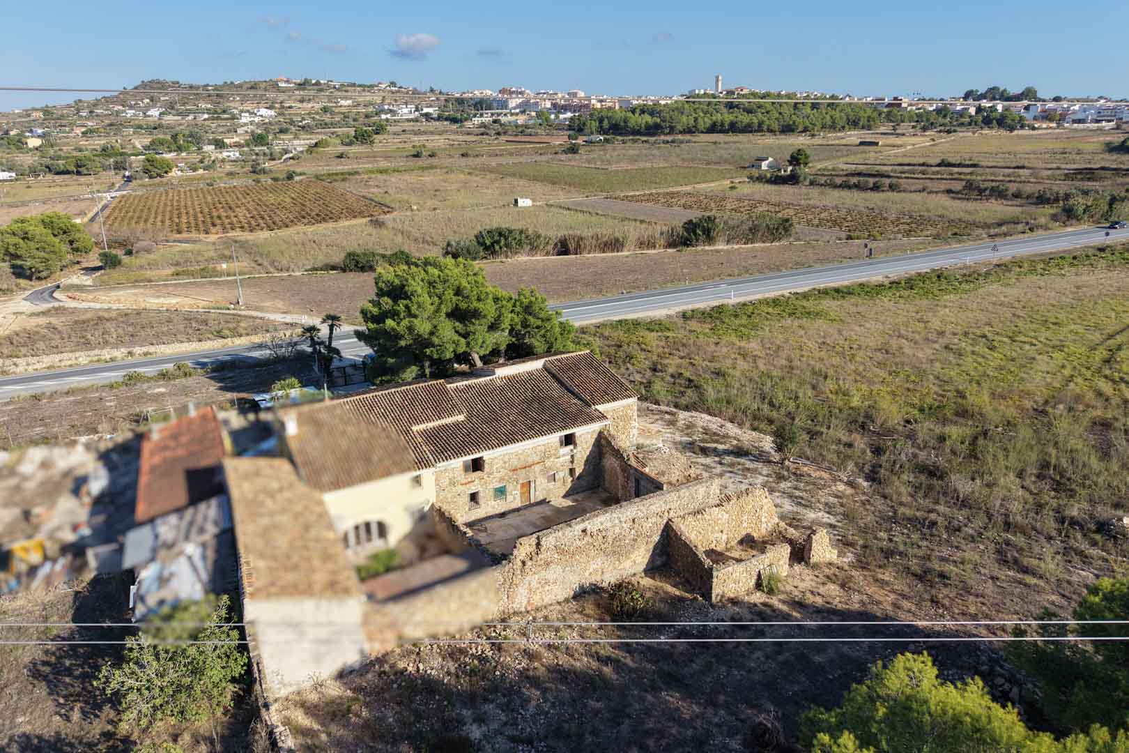 Vue aérienne de la maison traditionnelle en pierre et de ses annexes, située sur un vaste terrain rural avec vue sur le village de Teulada.