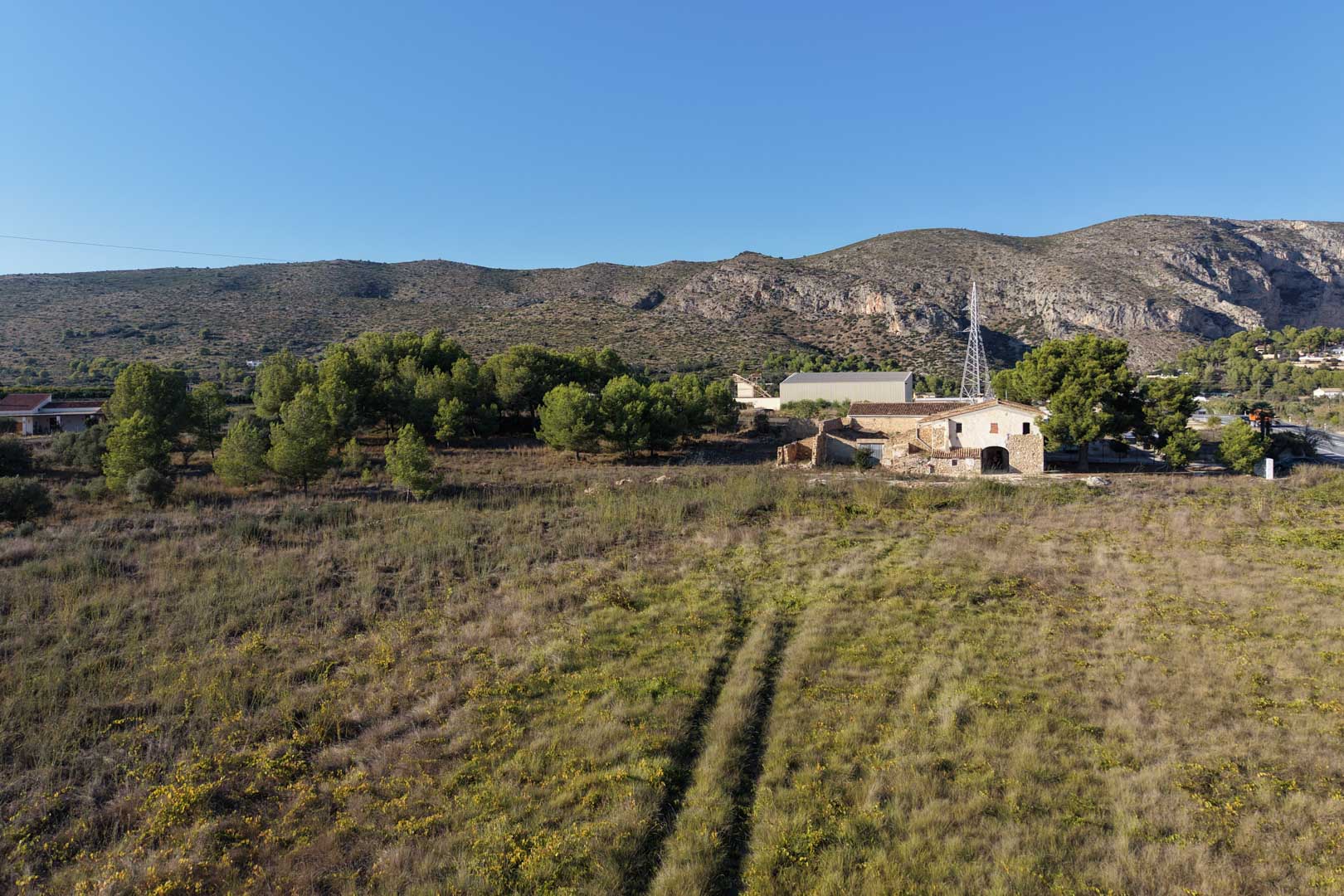Vue aérienne de la maison traditionnelle en pierre et de son vaste terrain plat, entourée de végétation méditerranéenne avec les montagnes de Teulada en arrière-plan.