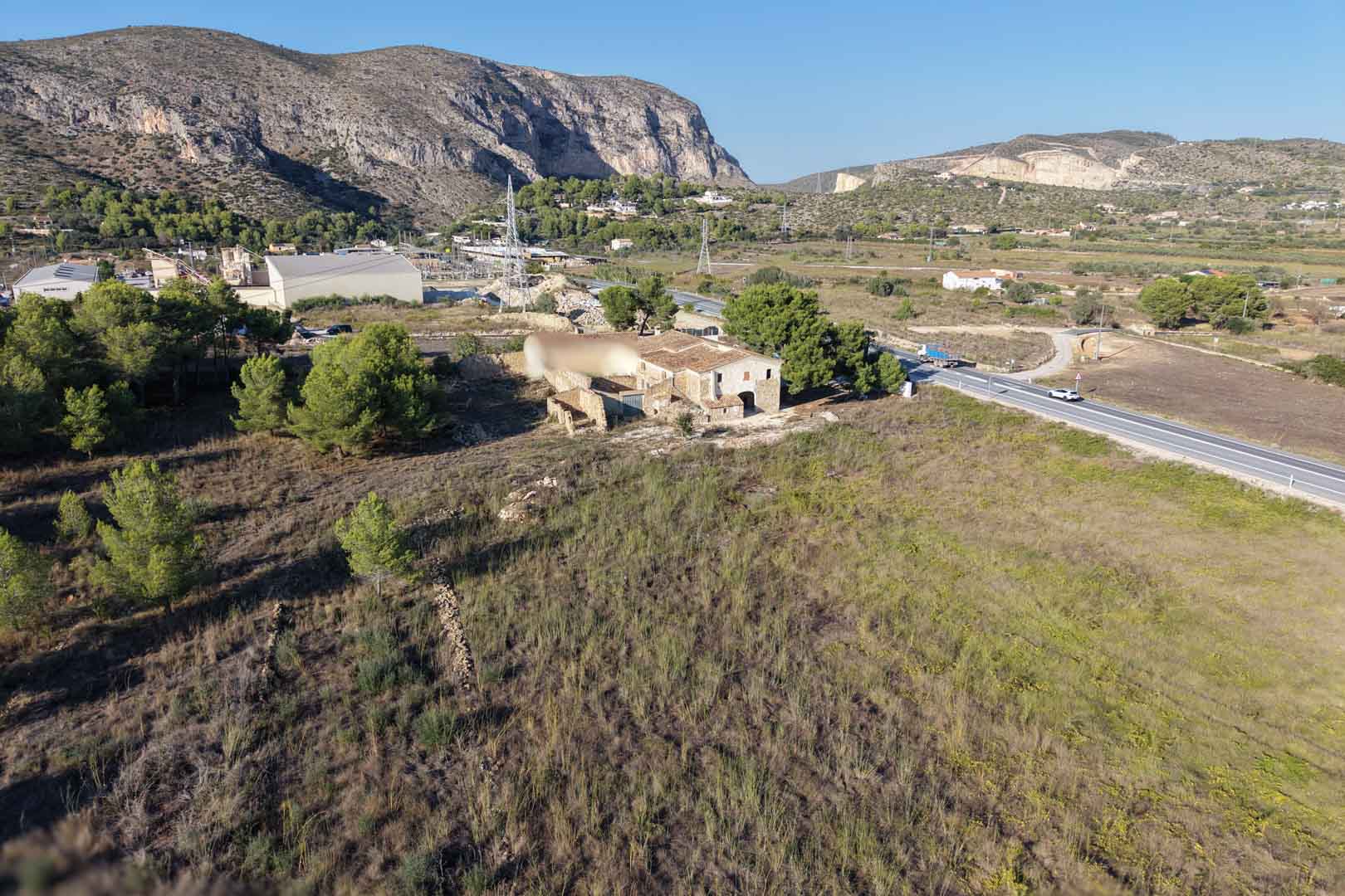 Vue aérienne de la maison traditionnelle en pierre sur une vaste parcelle plane à Teulada, avec les montagnes en arrière-plan et un accès routier.