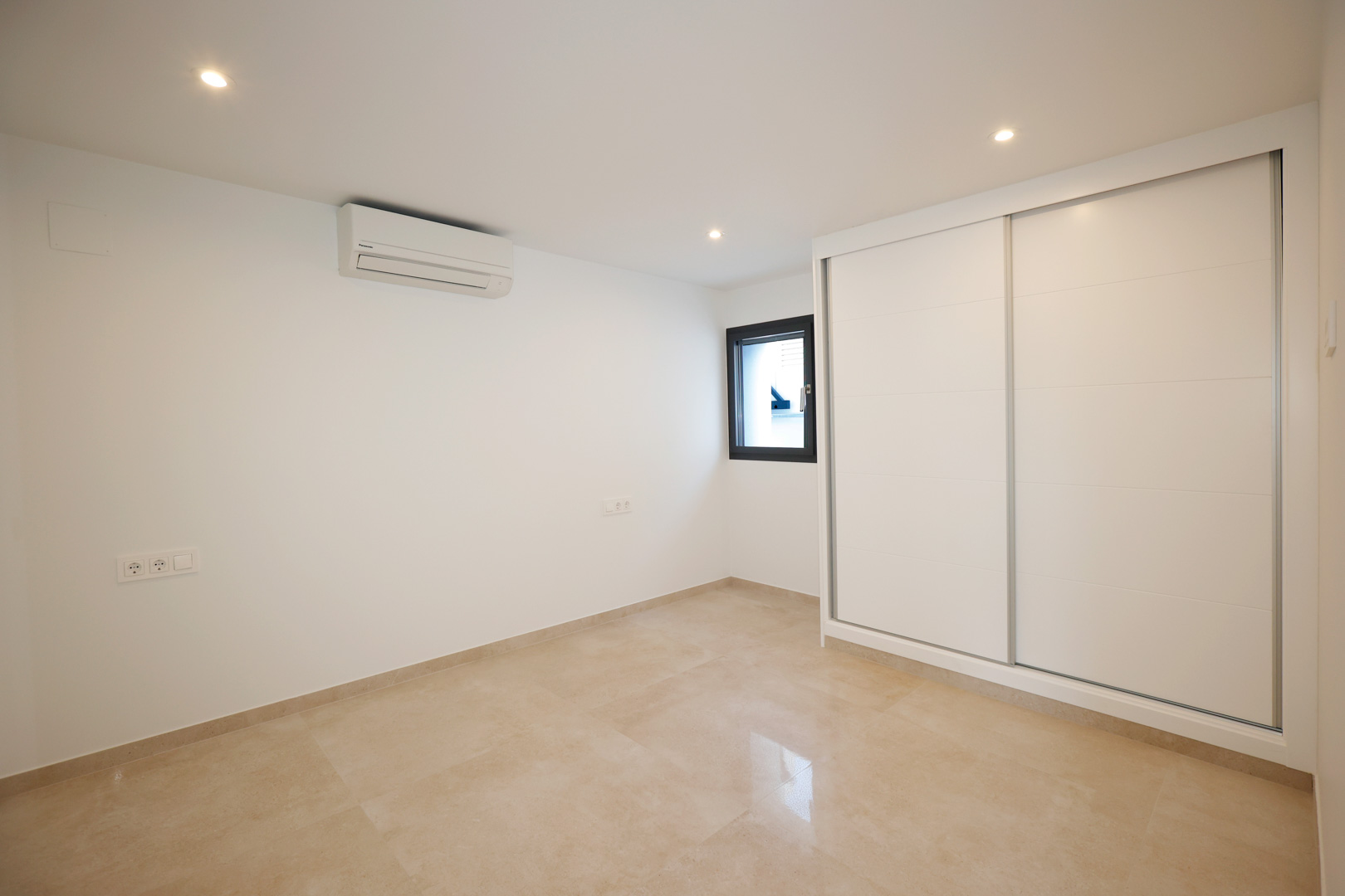 Chambre avec placard intégré à portes coulissantes blanches, sol en grès cérame, climatisation et spots LED encastrés au plafond.