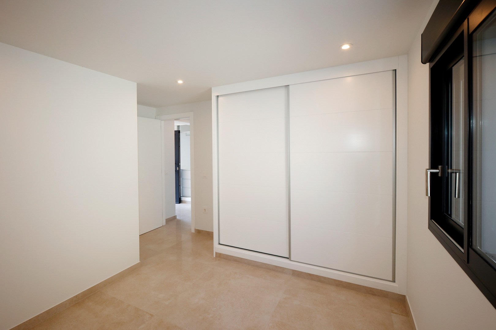 Chambre avec placard intégré à portes coulissantes blanches, sol en grès cérame et éclairage LED encastré près d'une fenêtre en aluminium noir.