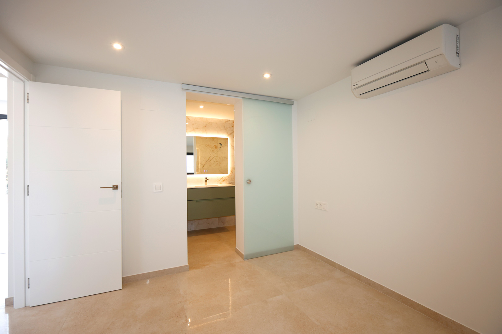 Chambre avec sol en grès cérame, climatisation et porte coulissante en verre dépoli ouvrant sur une salle de bain attenante moderne.