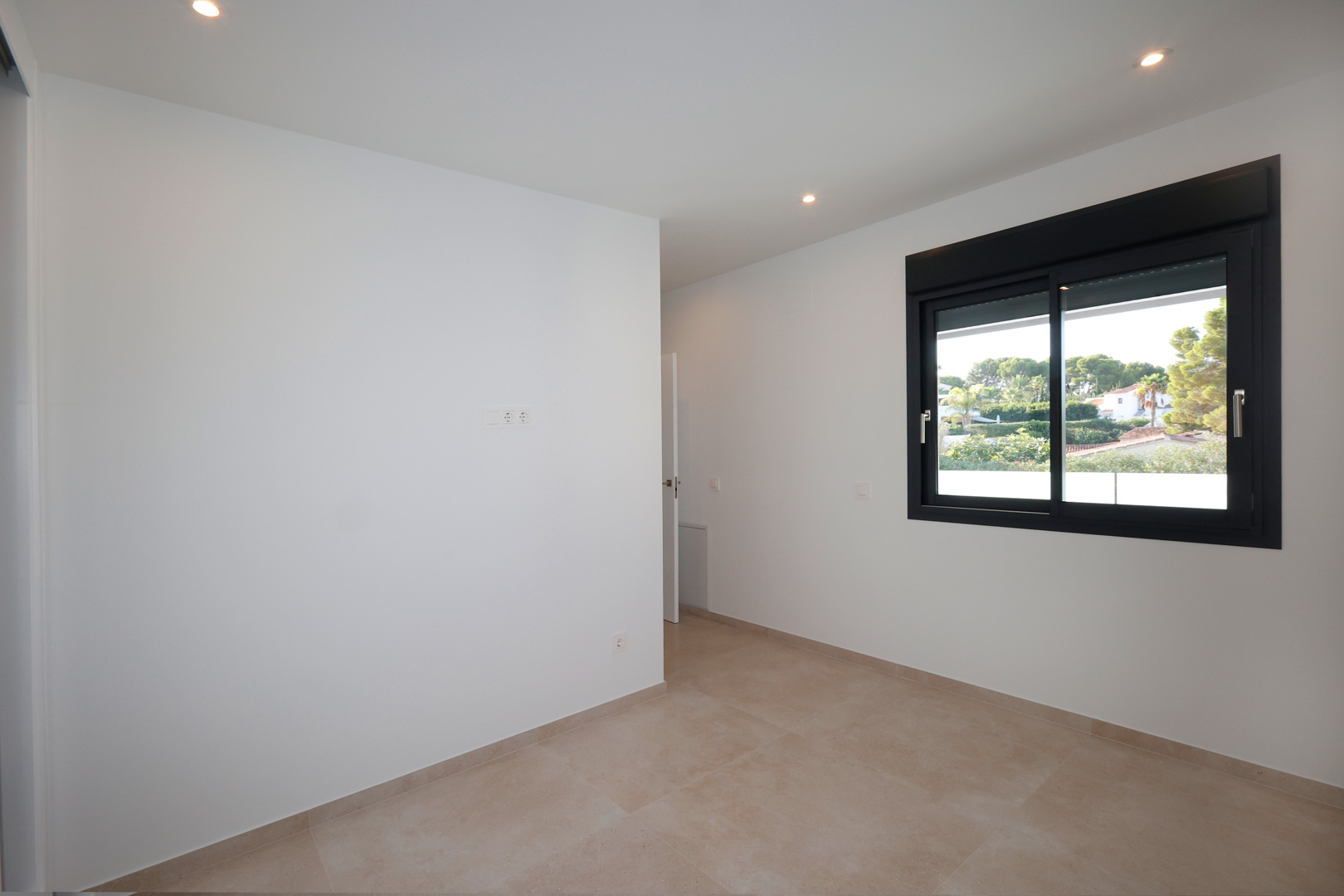 Chambre minimaliste avec murs blancs, sol en grès cérame et fenêtre en aluminium noir équipée de volets électriques offrant une vue sur la verdure.