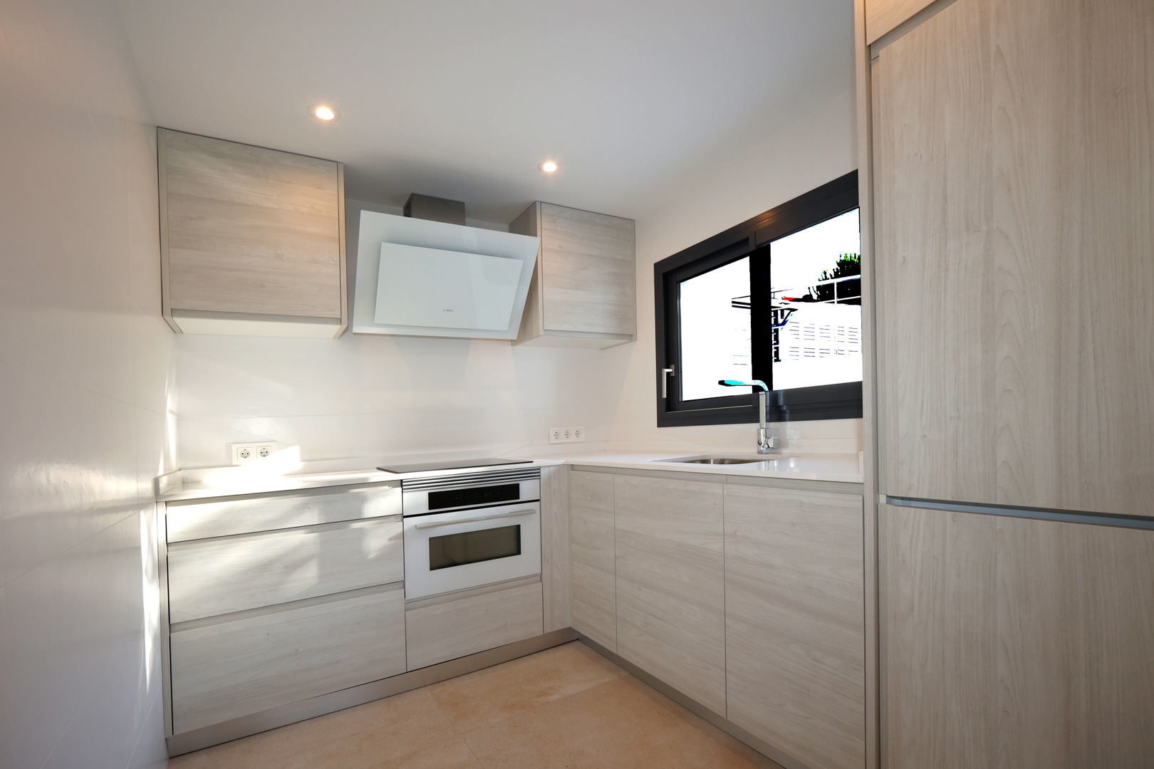 Cuisine fonctionnelle avec meubles en bois clair, plan de travail en Silestone blanc, plaque à induction, four Bosch et hotte décorative.