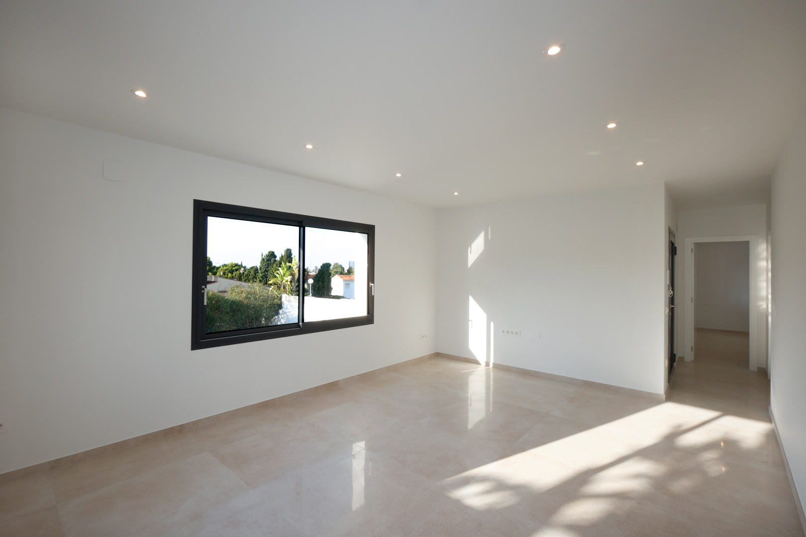 Espace de vie lumineux avec sol en grès cérame, éclairage LED encastré et fenêtre Technal. Le couloir mène à l'entrée équipée d'un système d'interphone.