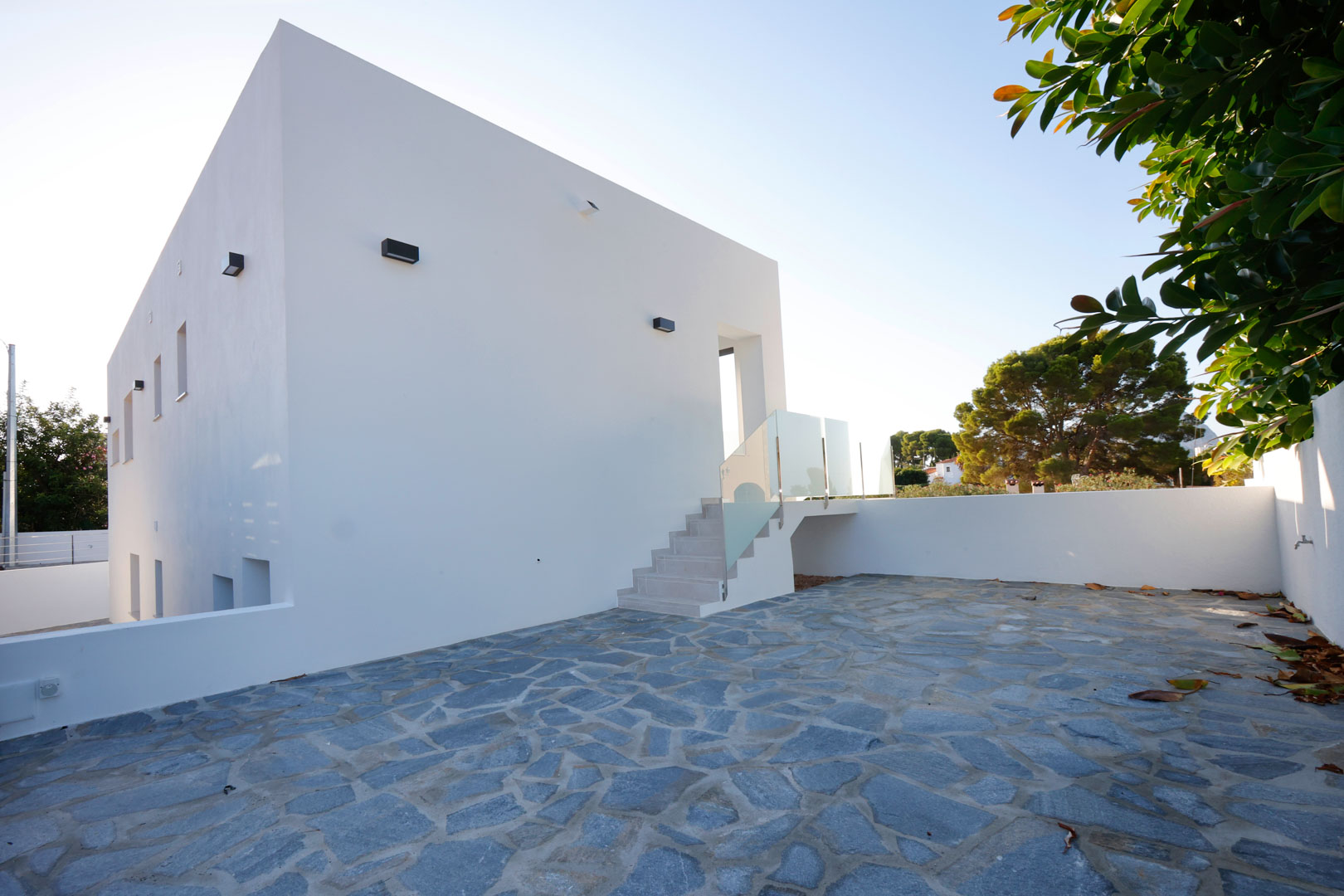 Façade blanche minimaliste avec un grand patio en pierre naturelle irrégulière et escalier extérieur muni de garde-corps en verre et inox.