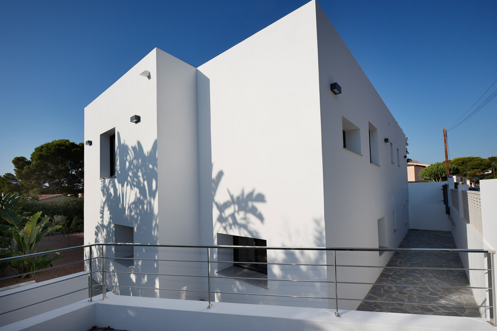 Façade minimaliste aux lignes épurées avec garde-corps en acier inoxydable et pavage en pierre naturelle. Les jeux d'ombres soulignent l'architecture contemporaine.