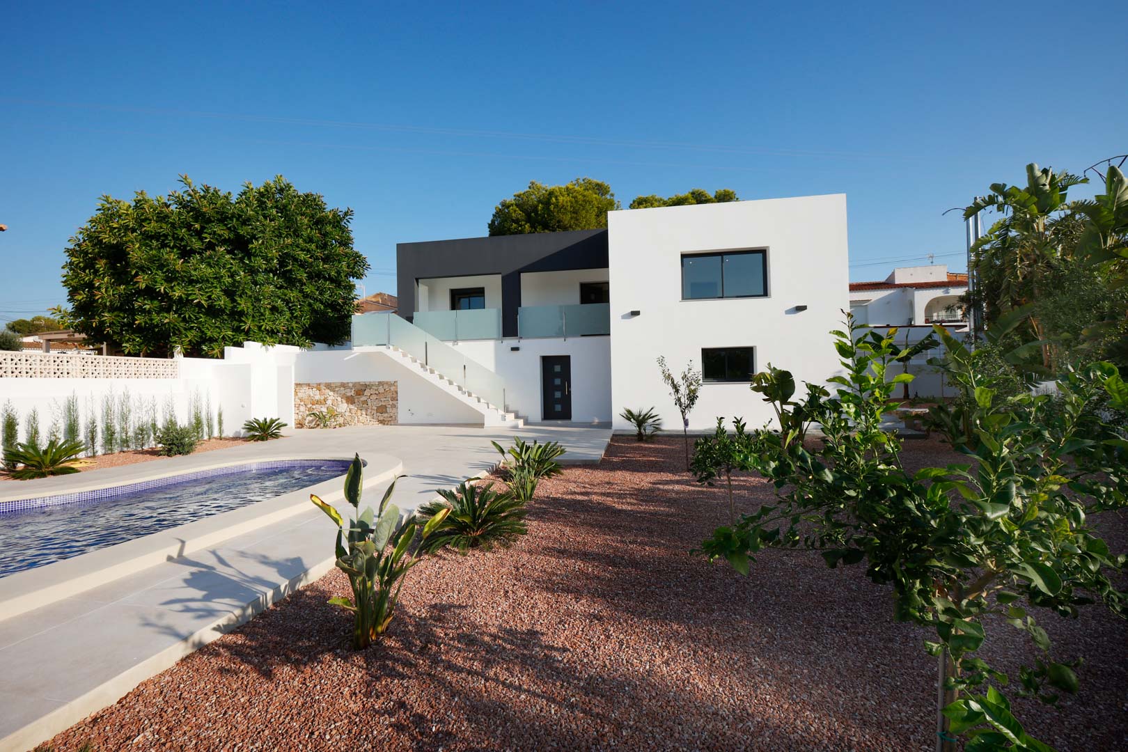 Façade moderne avec piscine, jardin en gravier de marbre rouge et escalier extérieur avec garde-corps en acier et verre. Comprend des murs en pierre et menuiserie Technal.