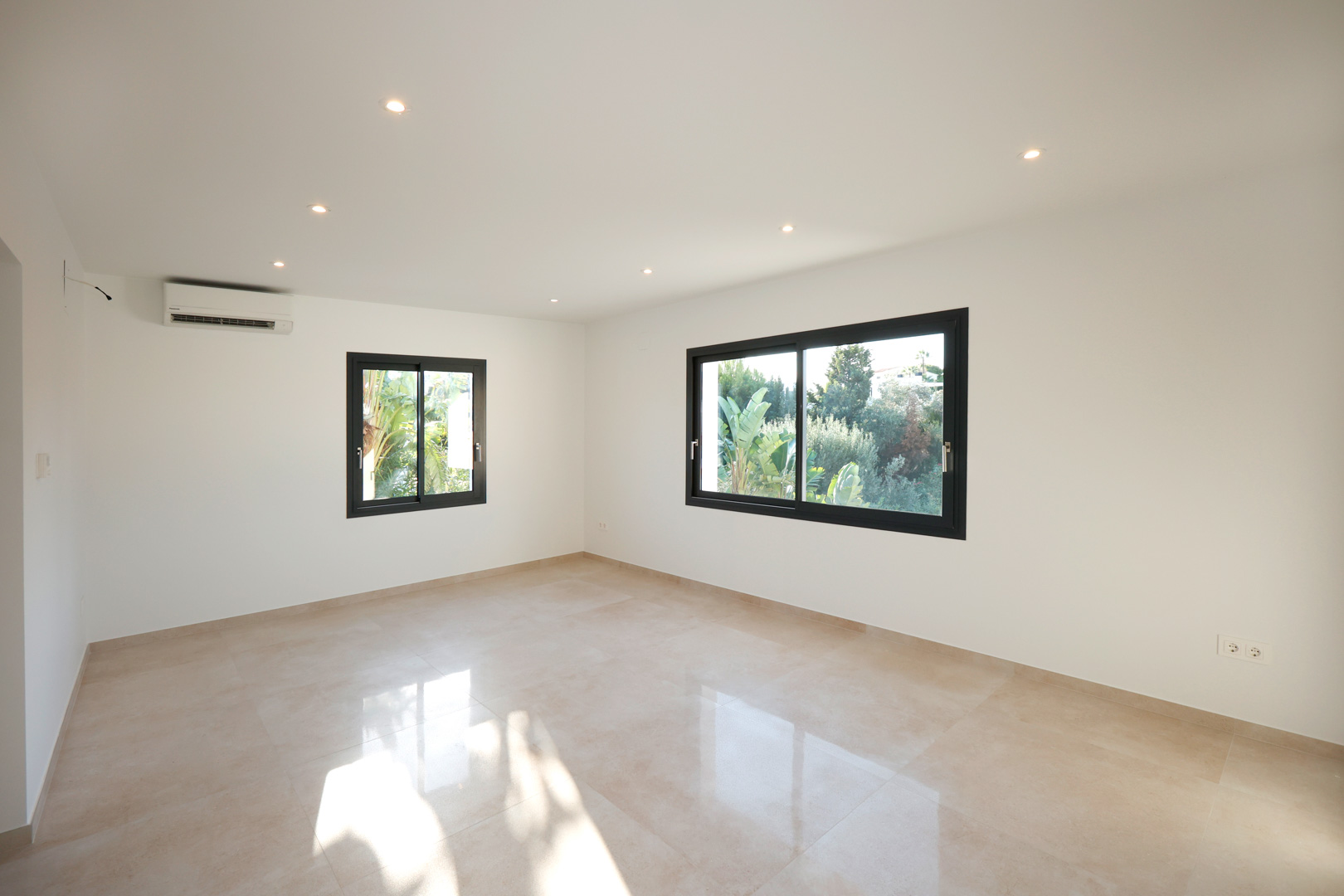 Pièce lumineuse avec sol en grès cérame, éclairage LED encastré, climatisation réversible et fenêtres en aluminium noir avec vue sur la végétation.