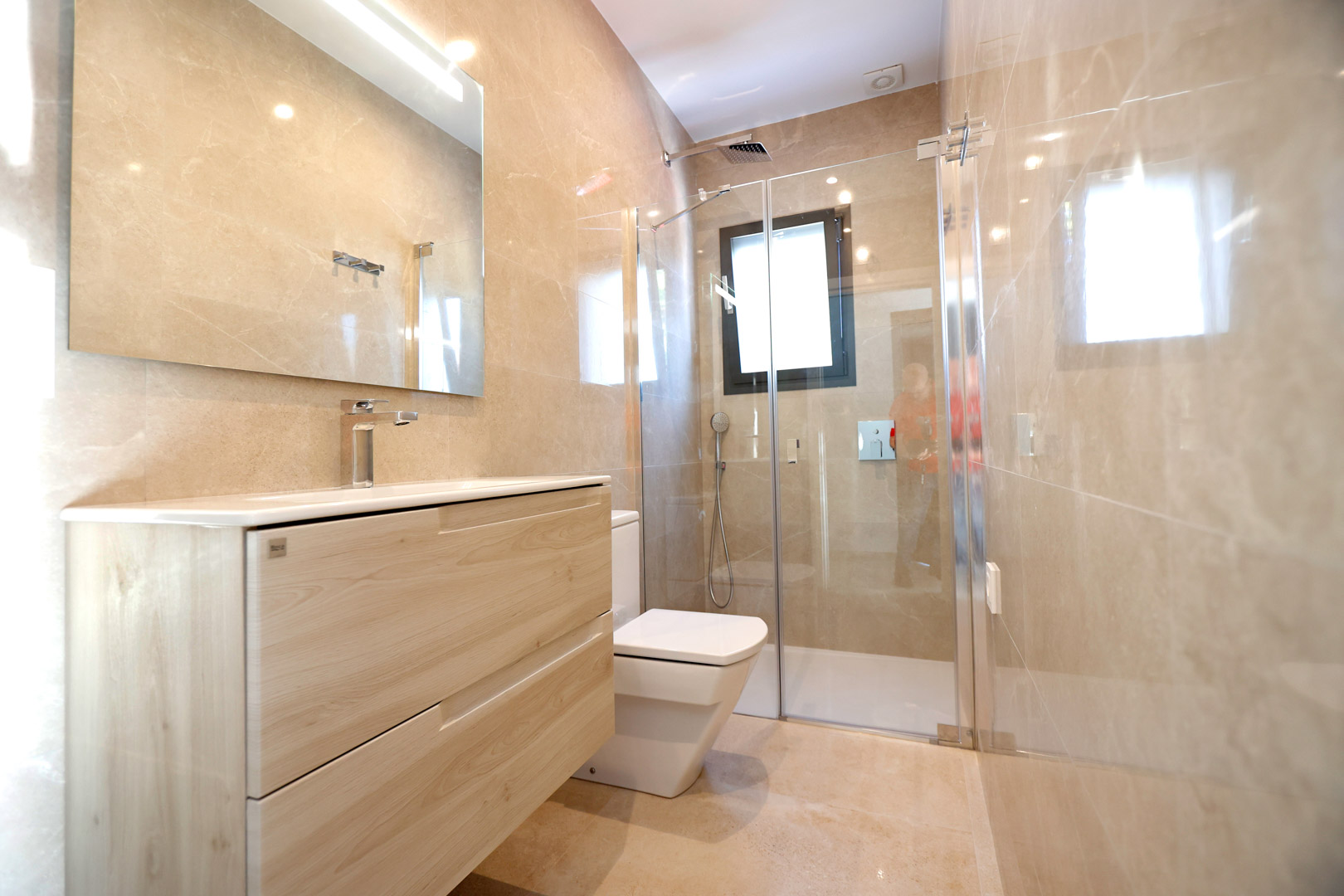 Salle de bain moderne avec meuble en bois clair, douche vitrée et murs en grès cérame poli. Équipée d'une robinetterie chromée et d'un éclairage LED intégré.
