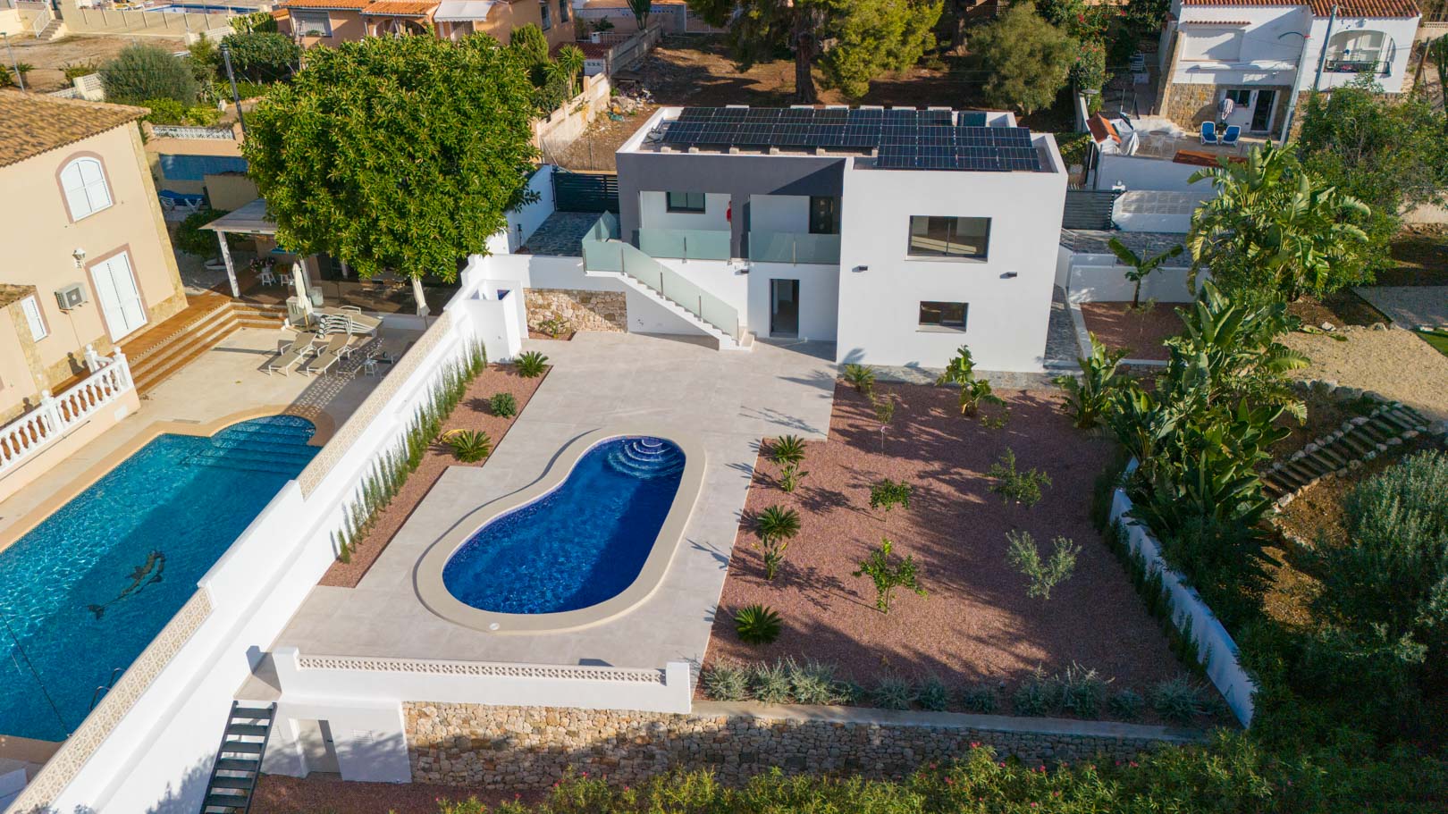 Vue aérienne de la villa avec piscine, jardin en gravier rouge et panneaux solaires sur le toit. On remarque les garde-corps en verre et les murs de soutènement en pierre naturelle.