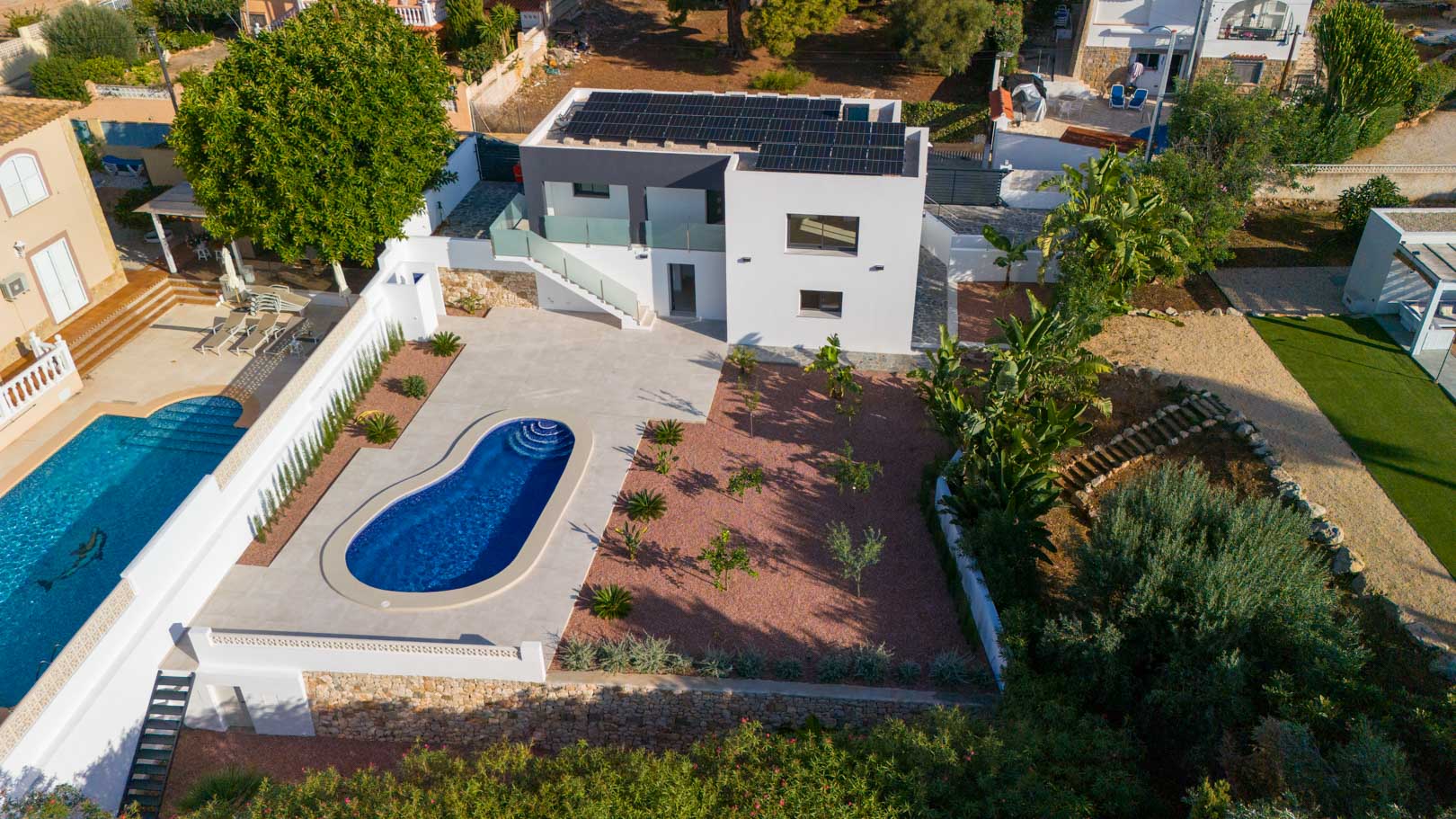 Vue aérienne de la villa moderne avec panneaux solaires, piscine de forme organique et jardin en gravier rouge bordé de murs en pierre naturelle.
