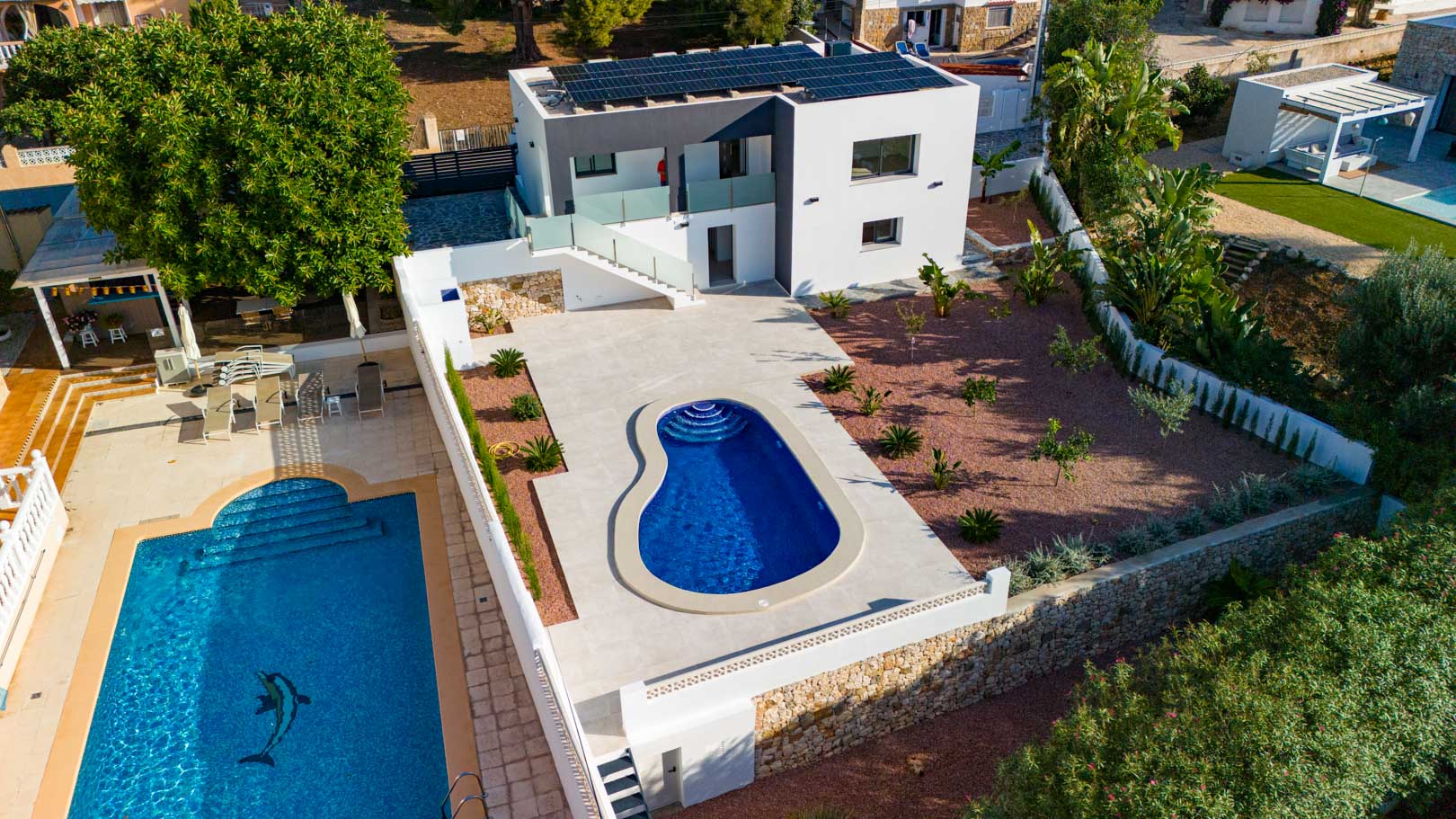 Vue aérienne de la villa moderne avec panneaux solaires, piscine de forme organique, jardin en gravier rouge et murs en pierre naturelle. Les garde-corps en verre soulignent son design.