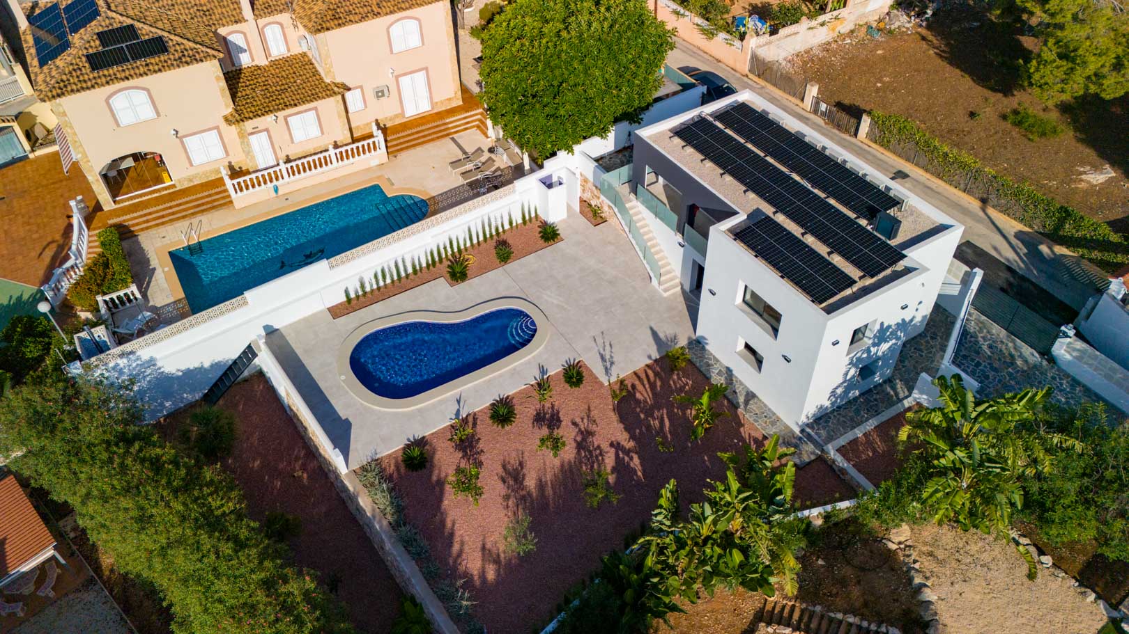 Vue aérienne de la villa moderne avec panneaux solaires, piscine en forme de rein et jardin en gravier rouge. Remarquez les garde-corps en verre et les murs en pierre.
