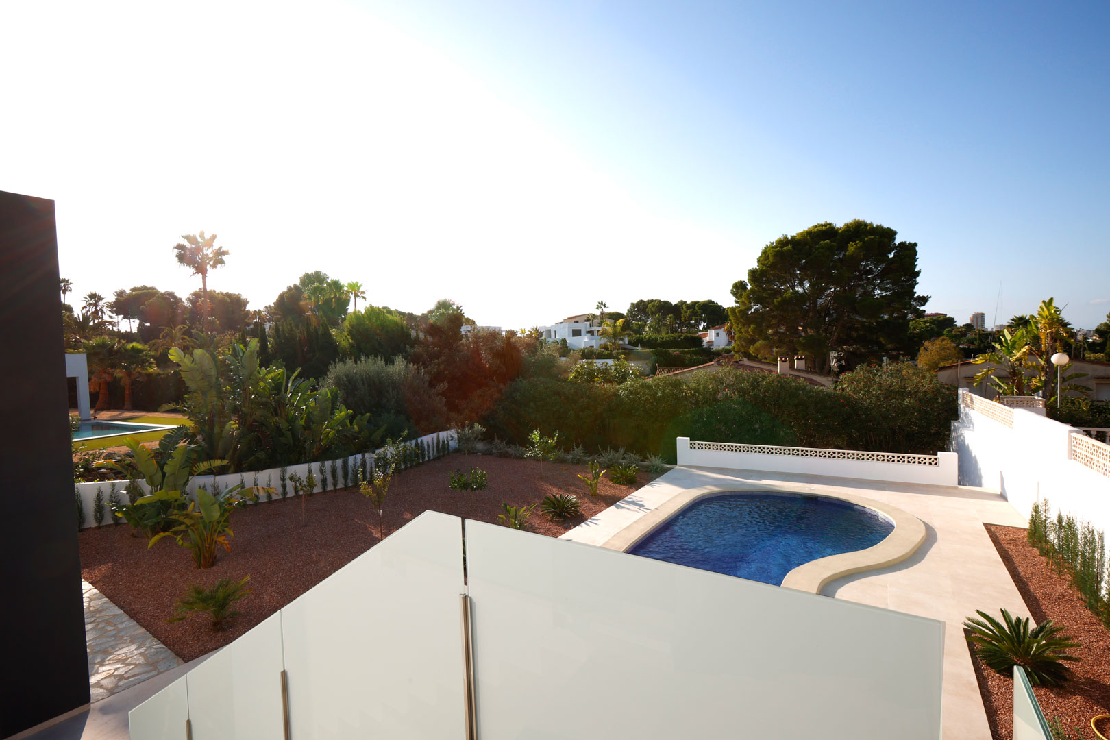 Vue depuis la terrasse supérieure avec garde-corps en verre, surplombant la piscine incurvée, le jardin en gravier rouge et le paysage méditerranéen.