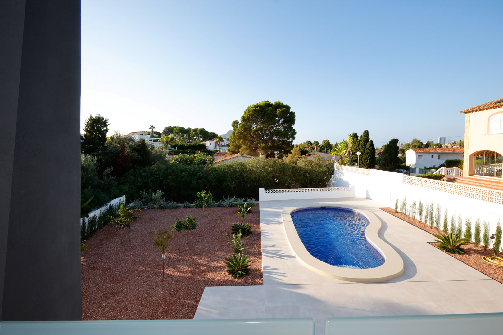 Vue surélevée du jardin avec piscine de forme organique, terrasse en grès blanc et zone de gravier de marbre rouge agrémentée de jeunes plantations.