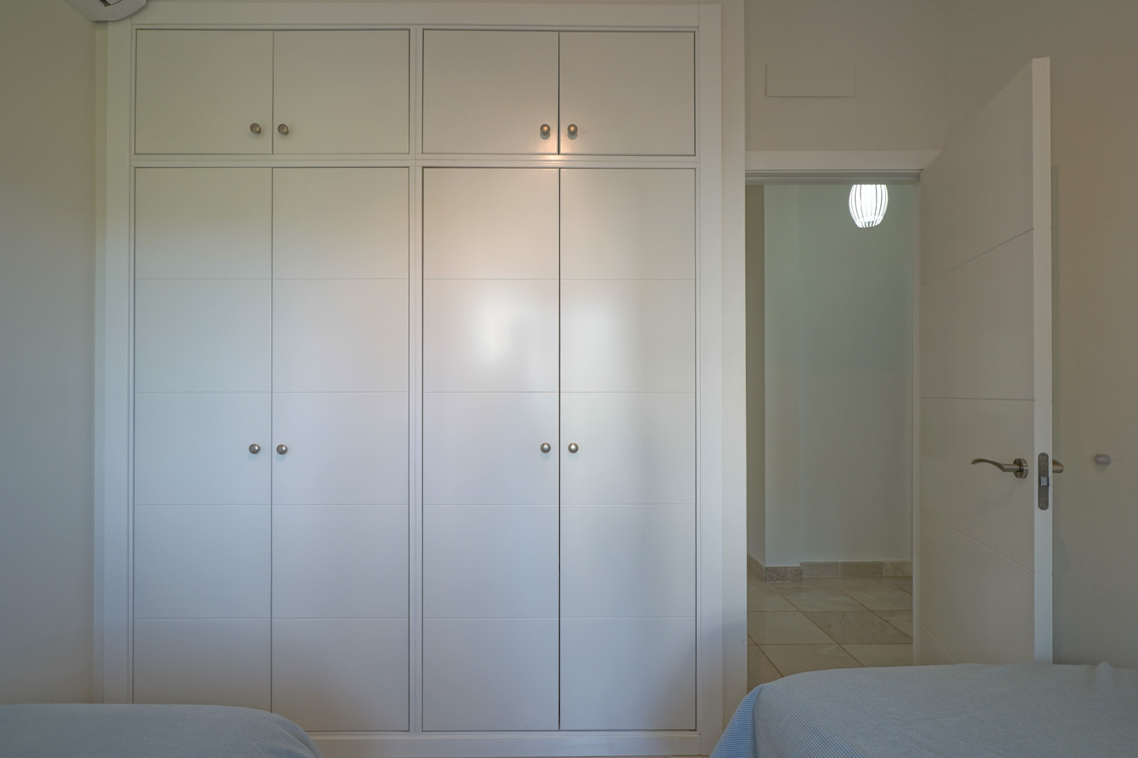 Armoire encastrée blanche avec poignées métalliques et rangements supérieurs, située à côté de la porte ouvrant sur un couloir au sol carrelé.