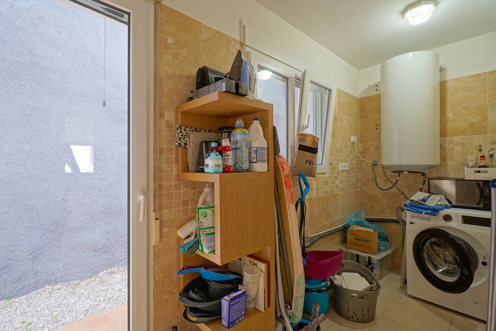 Buanderie fonctionnelle avec murs carrelés dans les tons crème, équipée d'un chauffe-eau électrique, d'un lave-linge, d'étagères en bois et d'un accès extérieur.