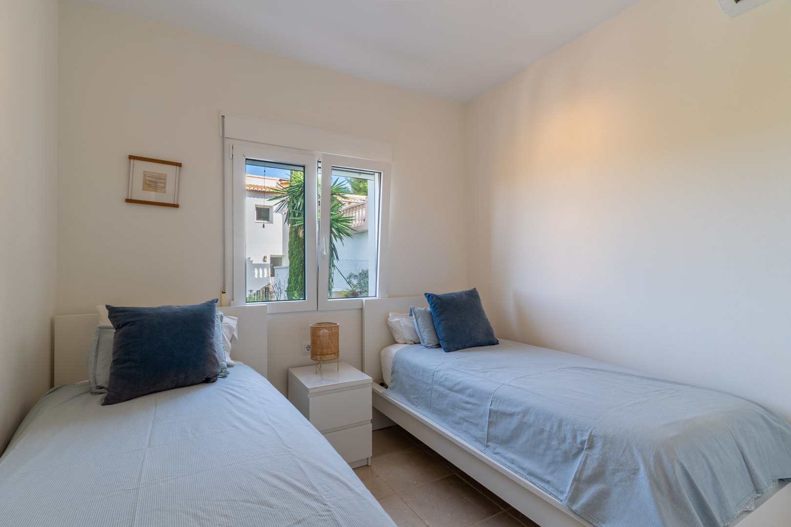 Chambre avec deux lits simples, murs blancs et fenêtre en PVC. L'espace est complété par une table de chevet et un éclairage naturel doux.
