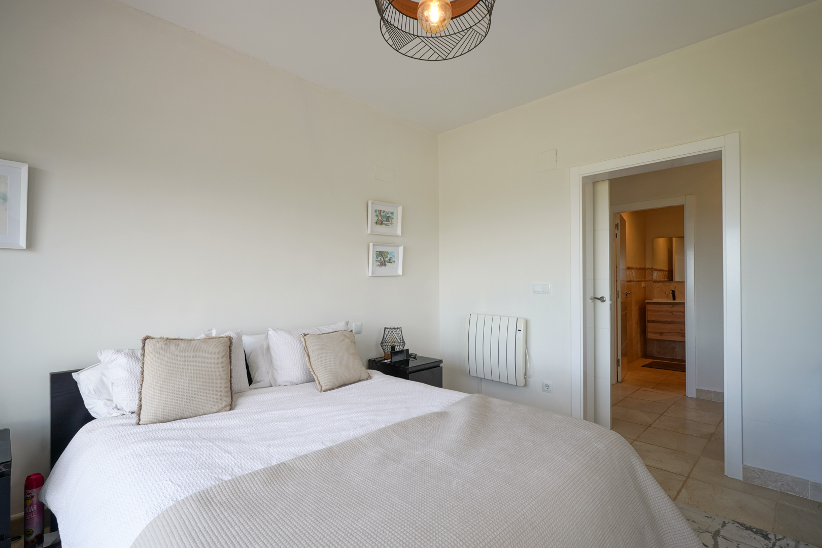 Chambre lumineuse avec sol en céramique, lit double et accès direct à une salle de bain attenante. Équipée d'un radiateur électrique et d'un luminaire design en métal.