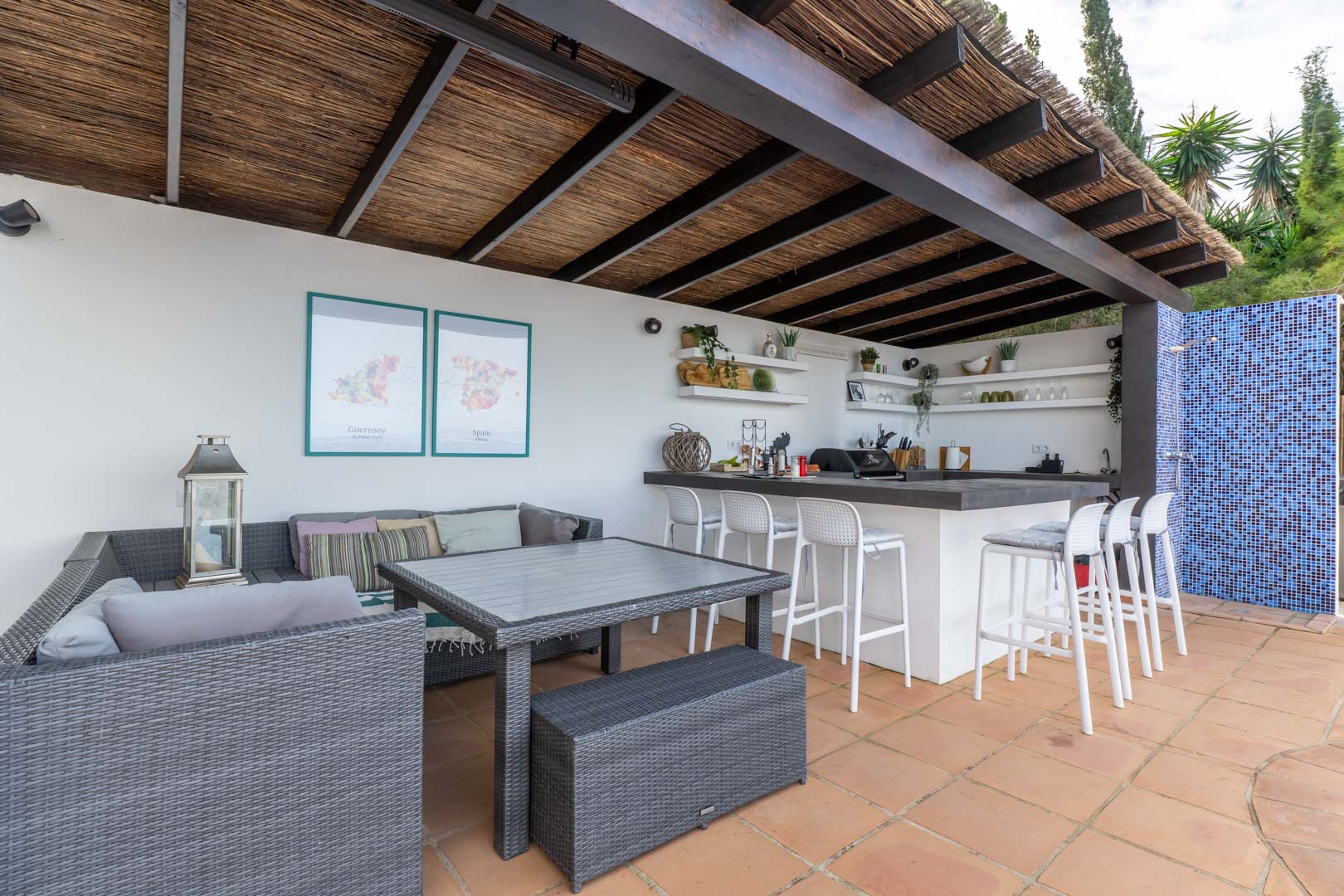 Cuisine d'été et salon extérieur sous une pergola en bois et canisse, comprenant un bar avec tabourets blancs, du mobilier en résine tressée et un sol en terre cuite.