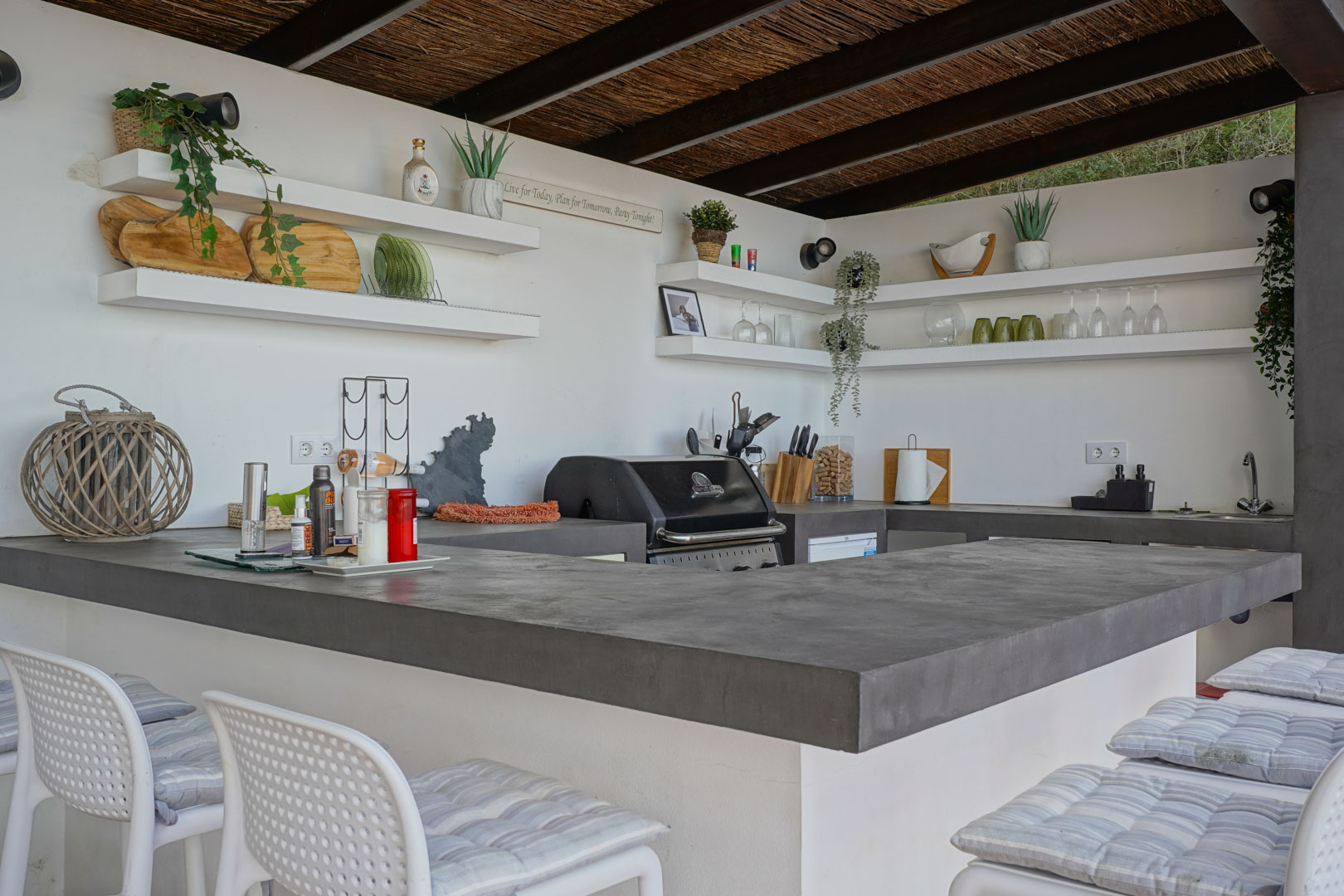 Cuisine d'été extérieure avec plans de travail en microciment gris, barbecue intégré, étagères murales et plafond en canisse avec poutres apparentes.