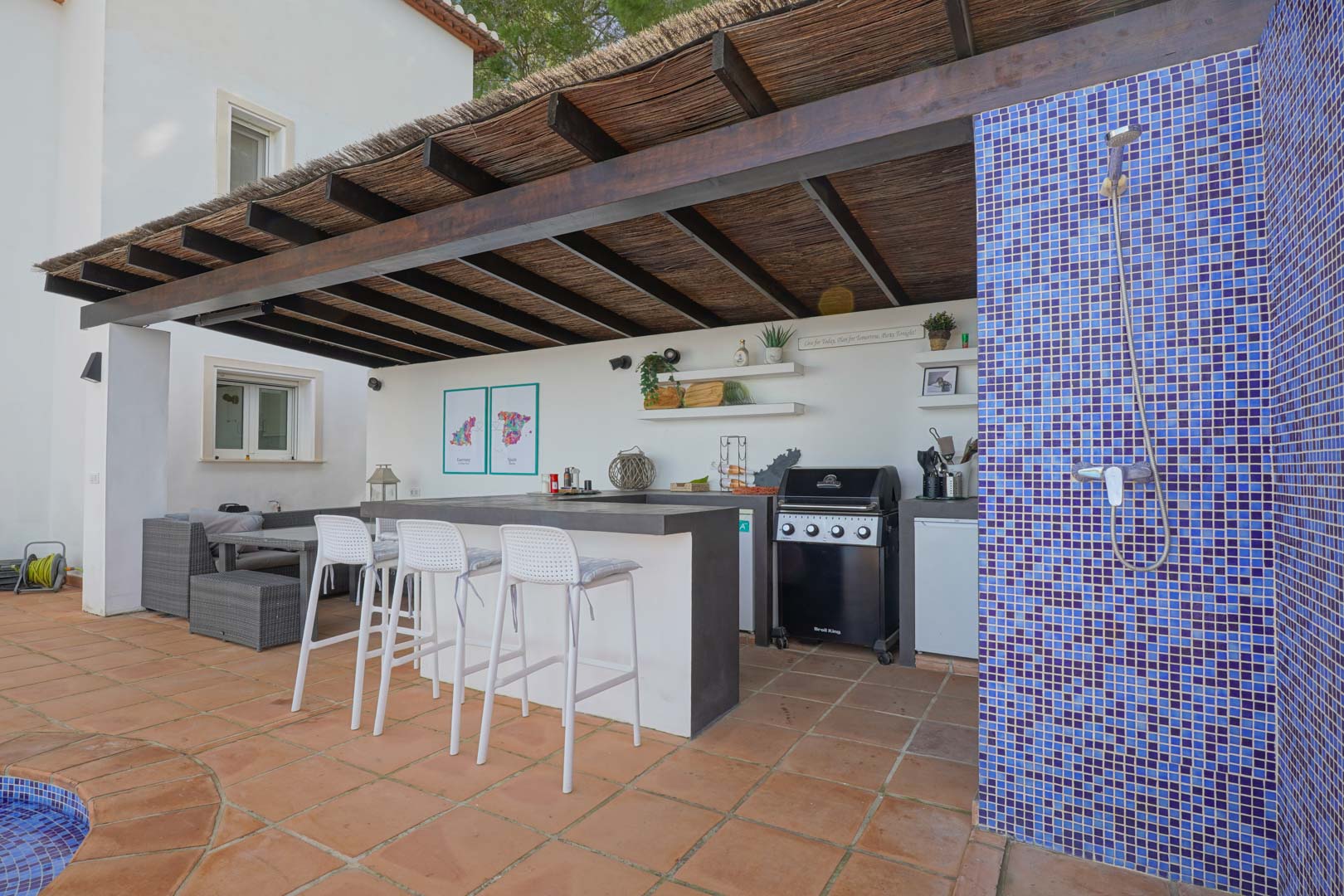 Cuisine d'été sous pergola en bois et brande, comprenant un barbecue à gaz, un bar avec tabourets blancs et une douche extérieure en mosaïque bleue.